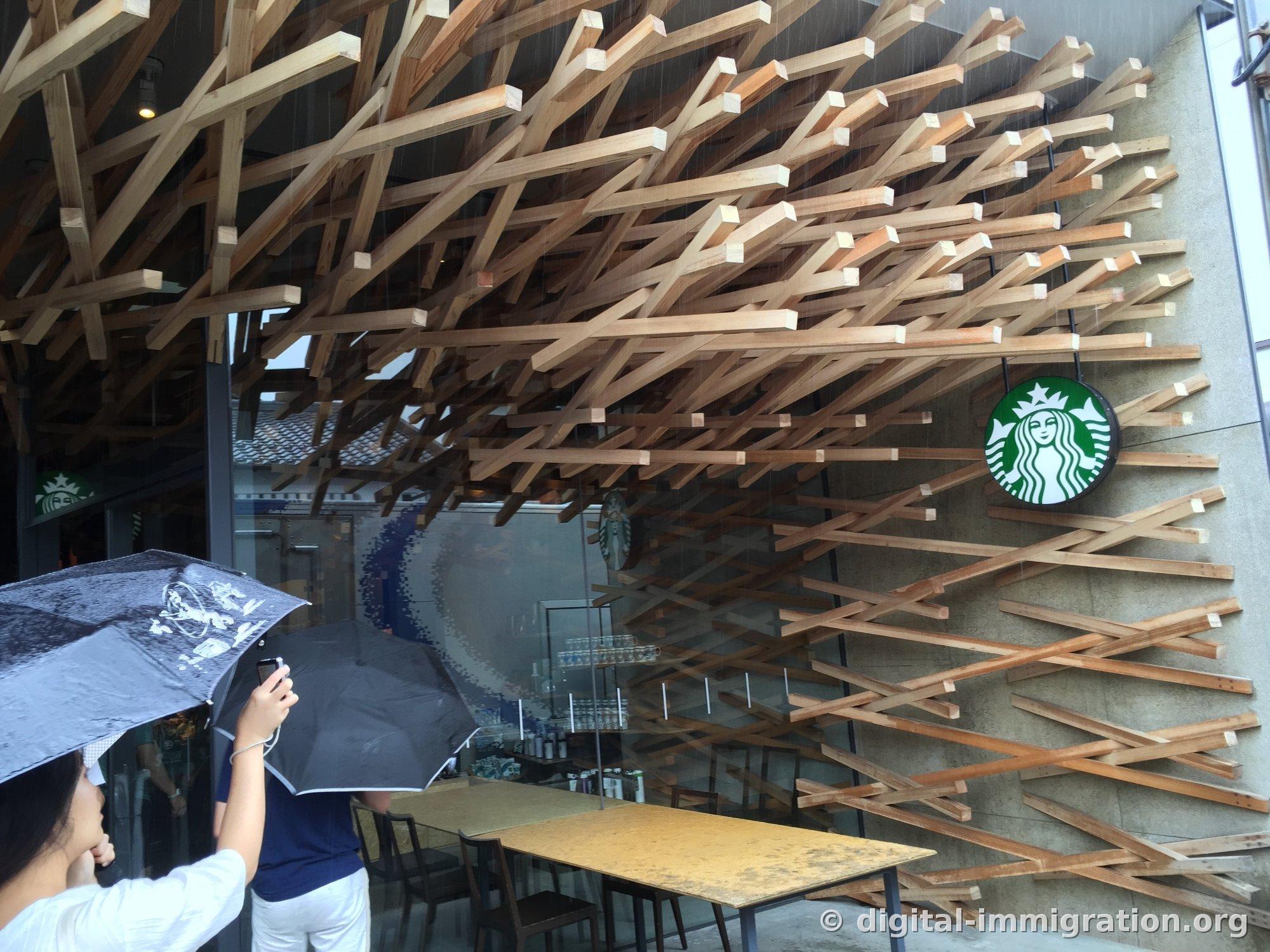 Interior wooden structure of Starbucks visible through glass with logo on wall, people outside with umbrellas taking photographs