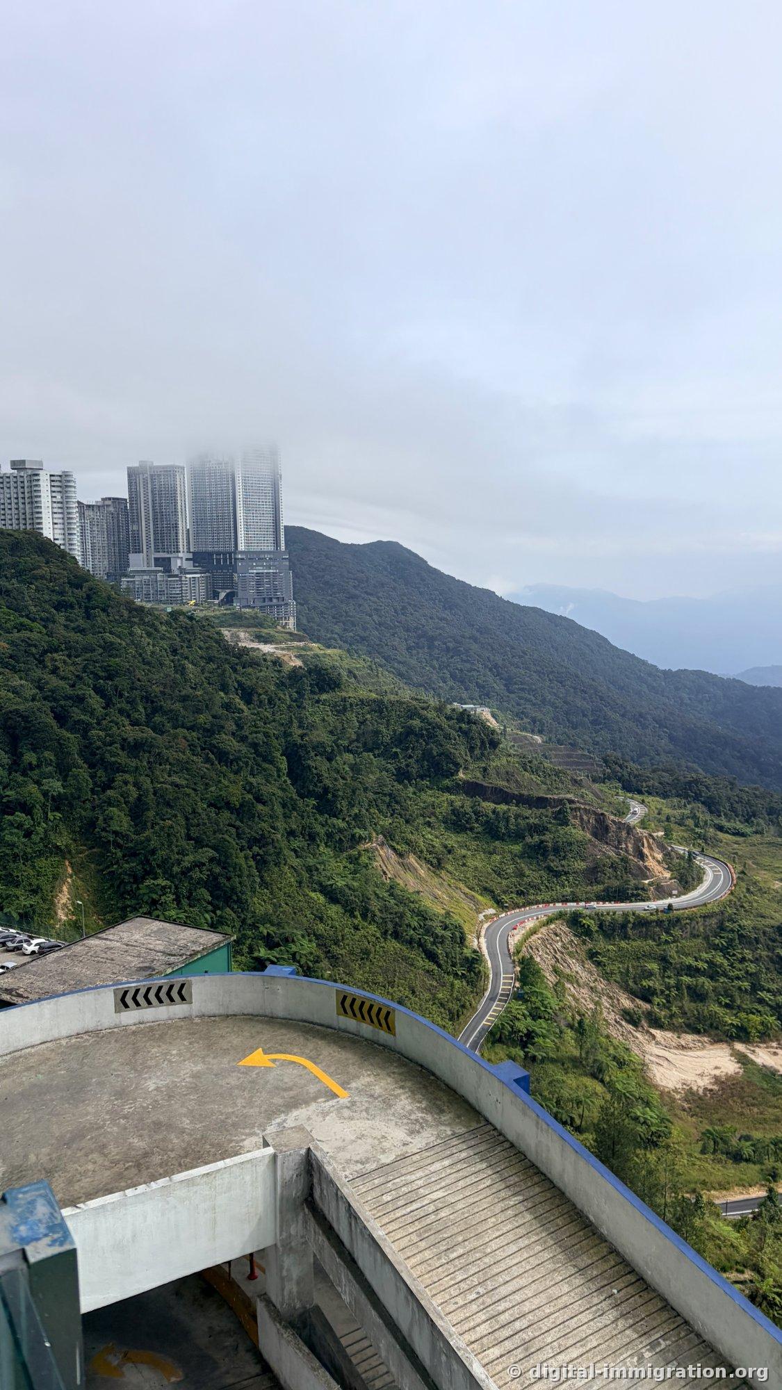 Overhead view from parking building showing winding mountain road around mountainside and apartment complex with peak cut off by clouds