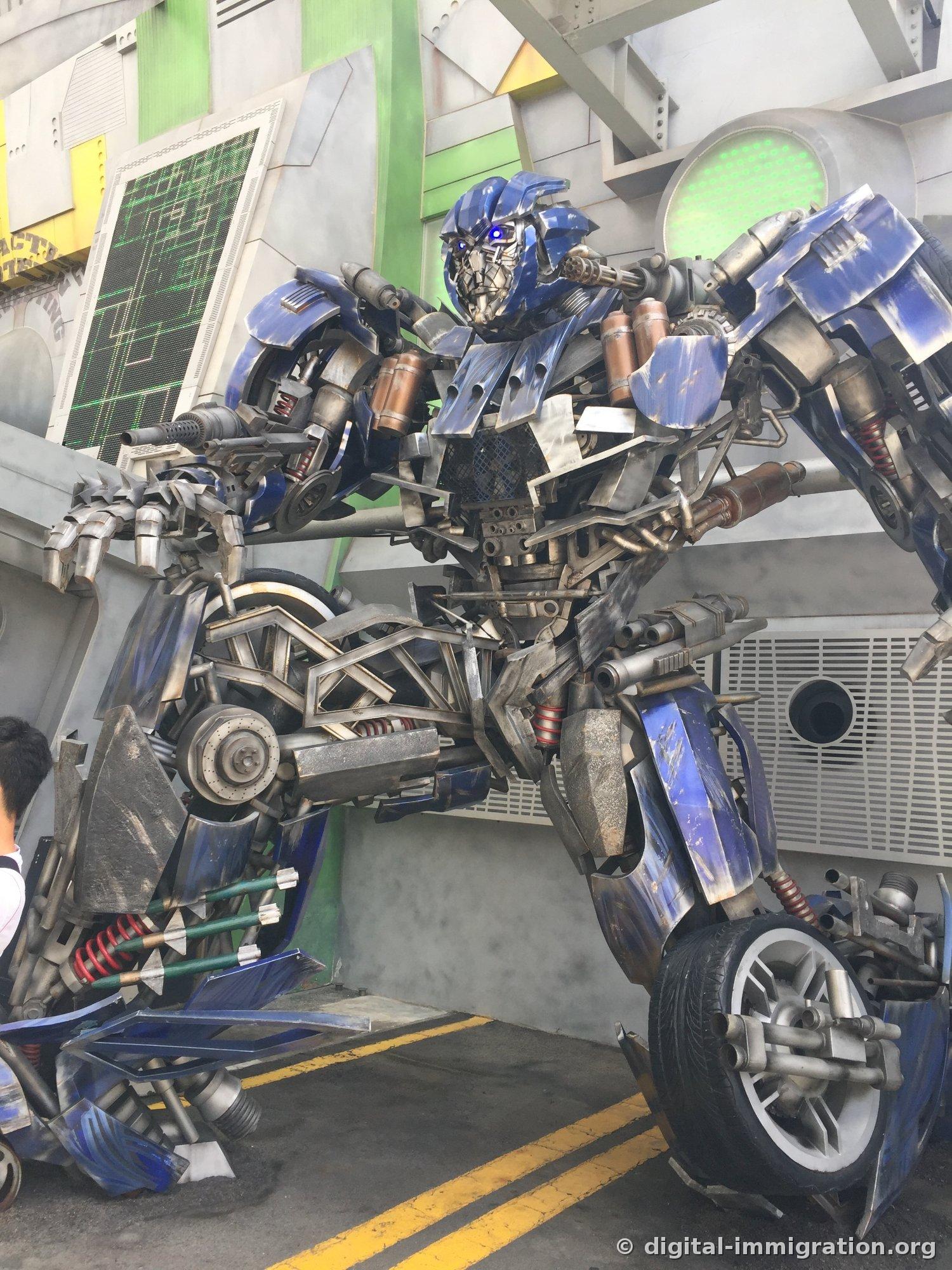 Blue Transformer Autobot sculpture assembled from car parts standing in a crouched position against the wall of an indoor corridor