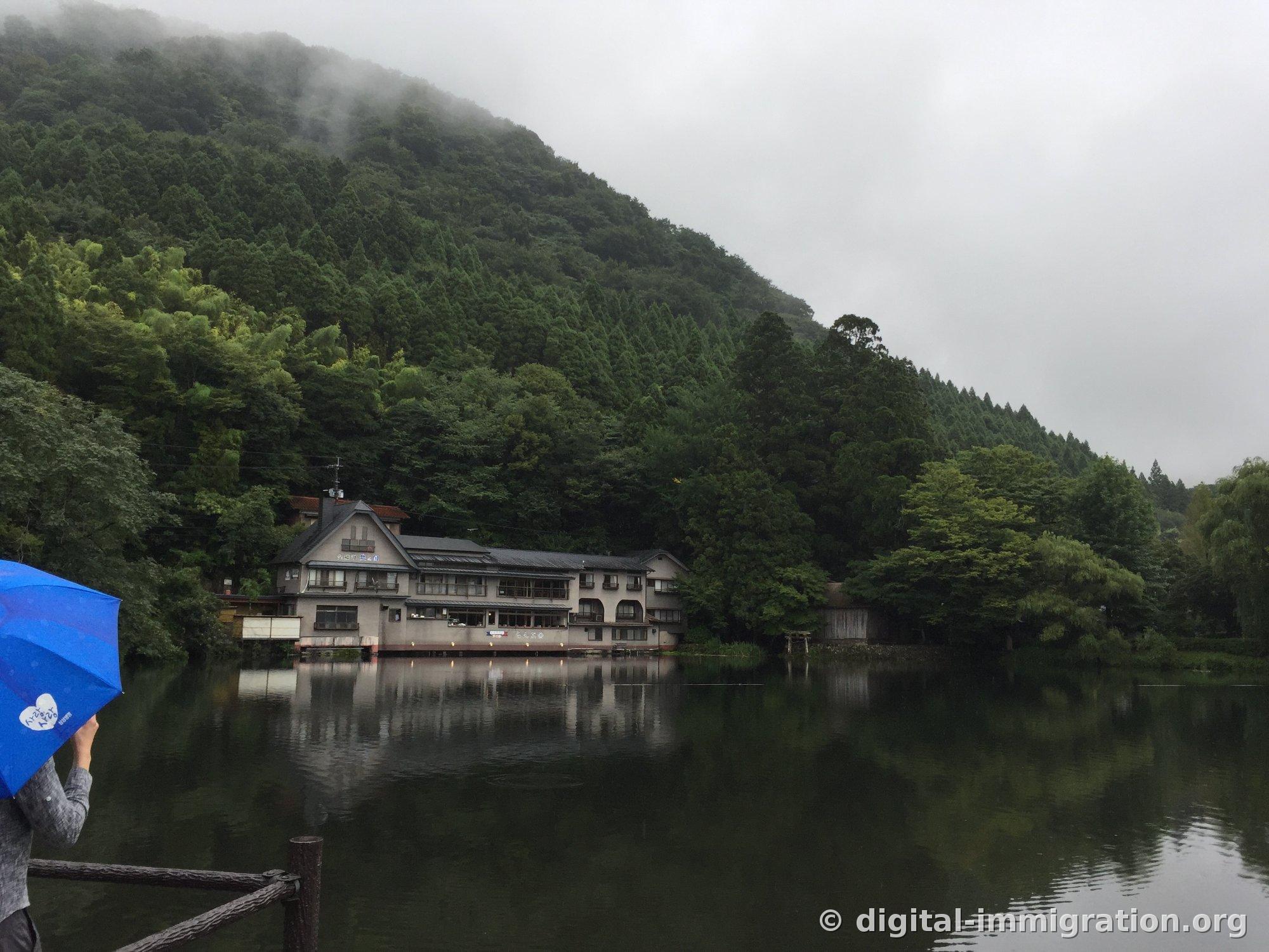 Une personne tenant un parapluie bleu regardant la montagne enveloppée de brouillard et les bâtiments de ryokan au bord du lac depuis la terrasse du lac Kinrin