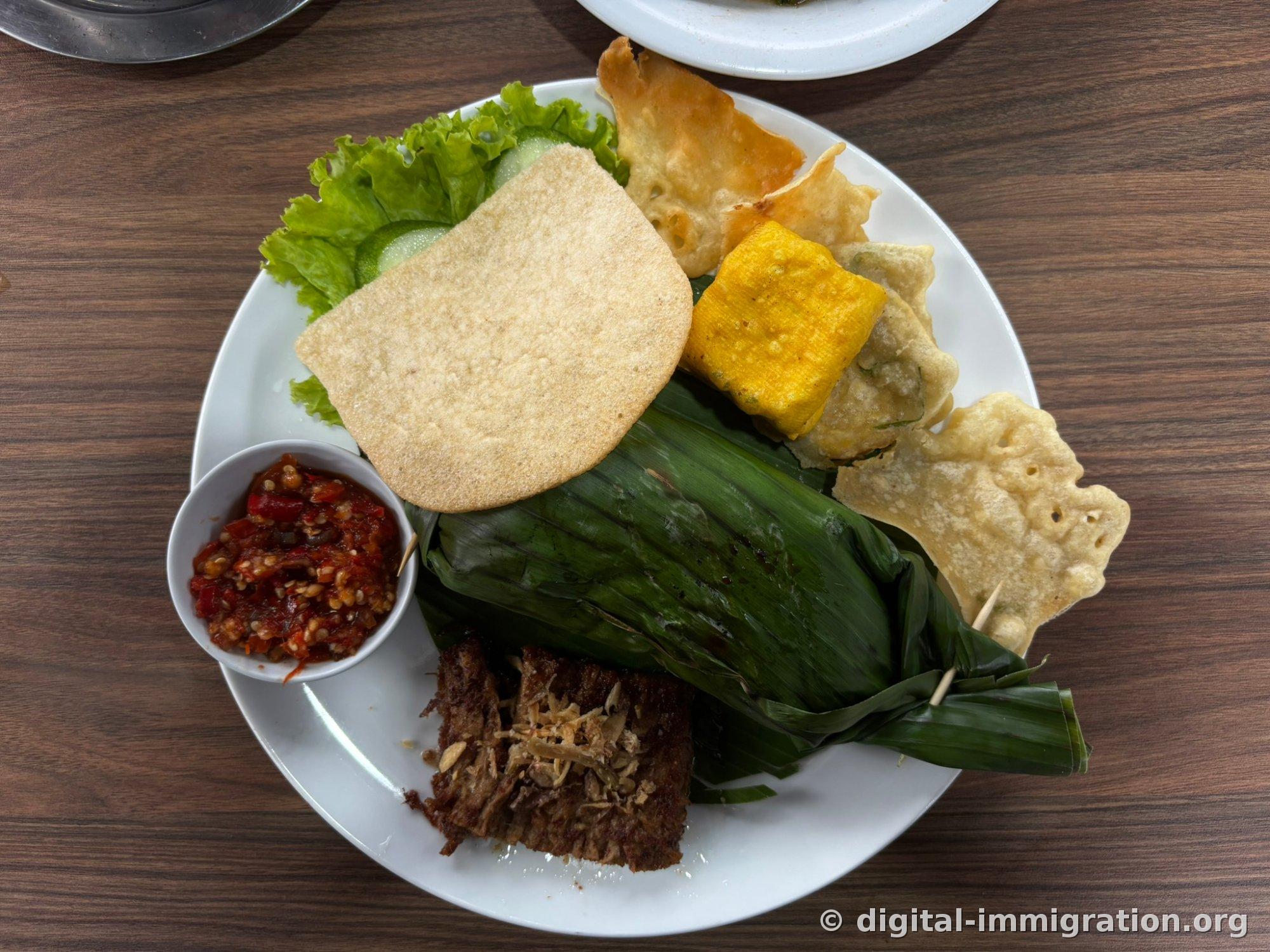 A full plate with banana-leaf wrapped grilled rice, large krupeuk, yellow fried tofu, braised beef, red sambal bowl, and lettuce and cucumber
