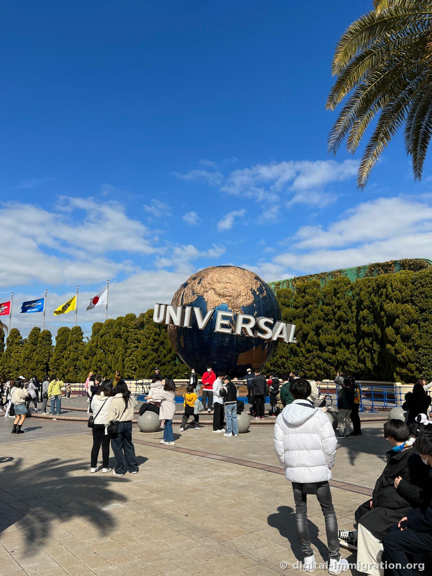 Spinning Universal globe monument against a blue sky with visitors taking photos in front, Japanese flag and Minions flags on the left
