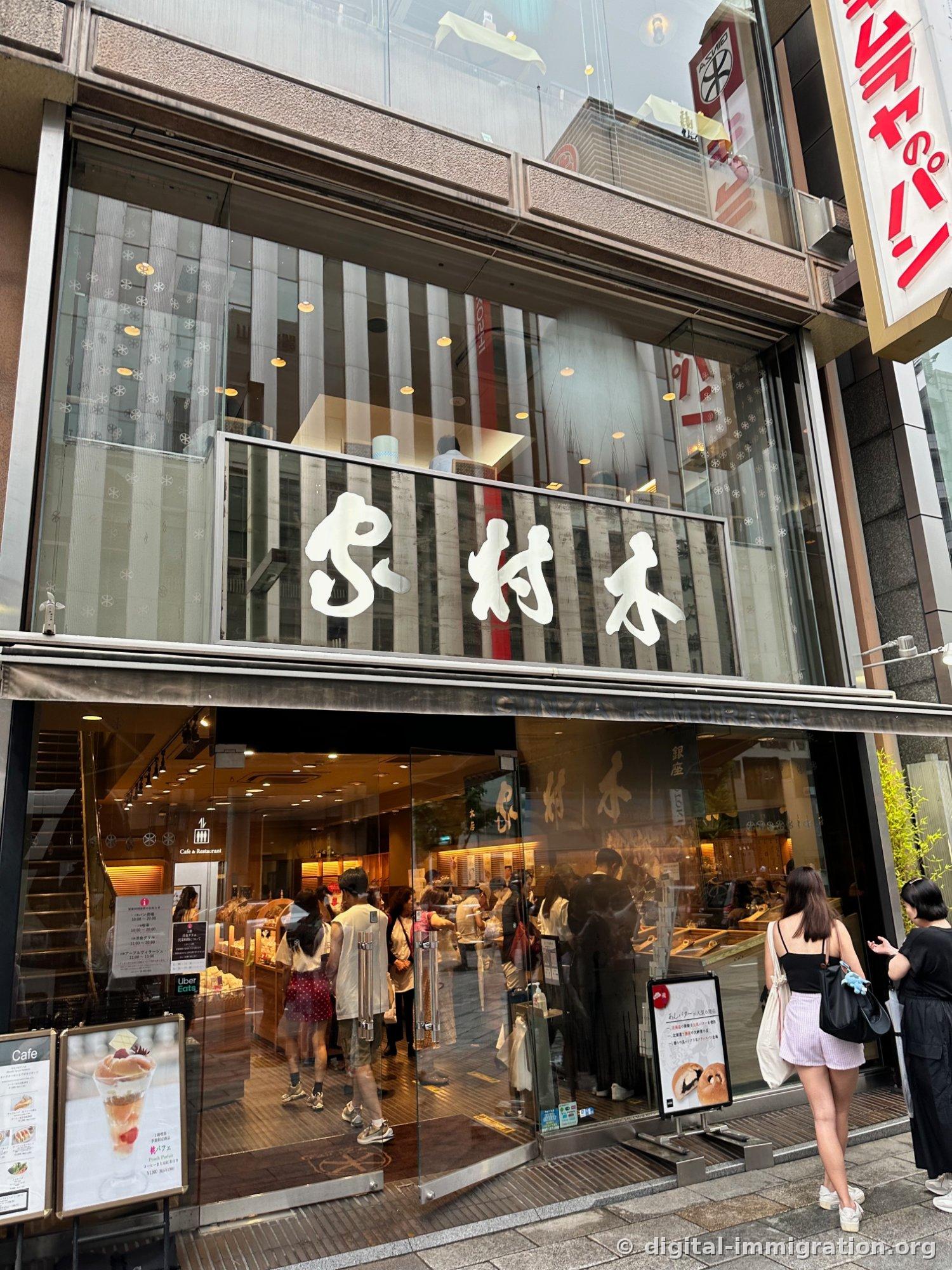 First-floor shop of Ginza Kimuraya with cursive 'Kimuraya' signboard inside glass façade, summer-dressed customers selecting bread