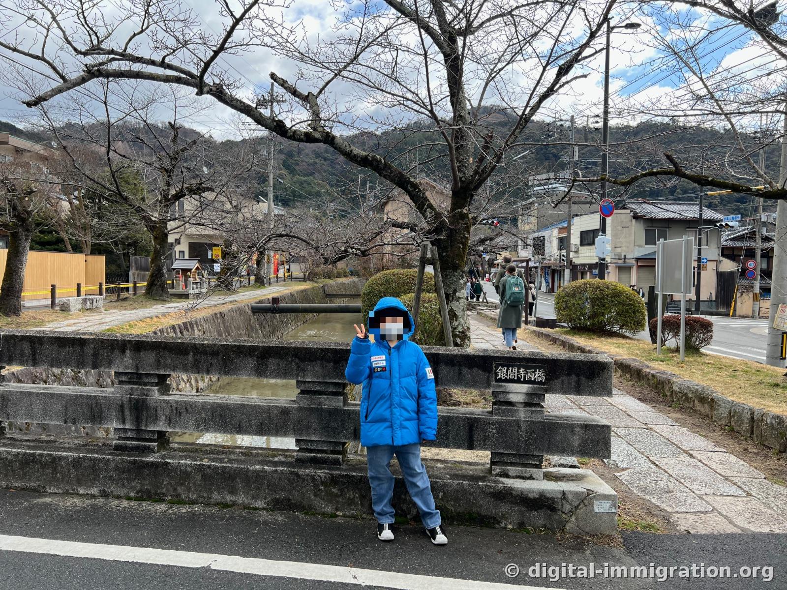 銀閣寺西橋라고 새겨진 돌난간 앞에 파란 패딩을 입은 아이가 서 있고, 뒤로 잎이 다 진 벚나무와 수로가 이어지는 겨울 풍경