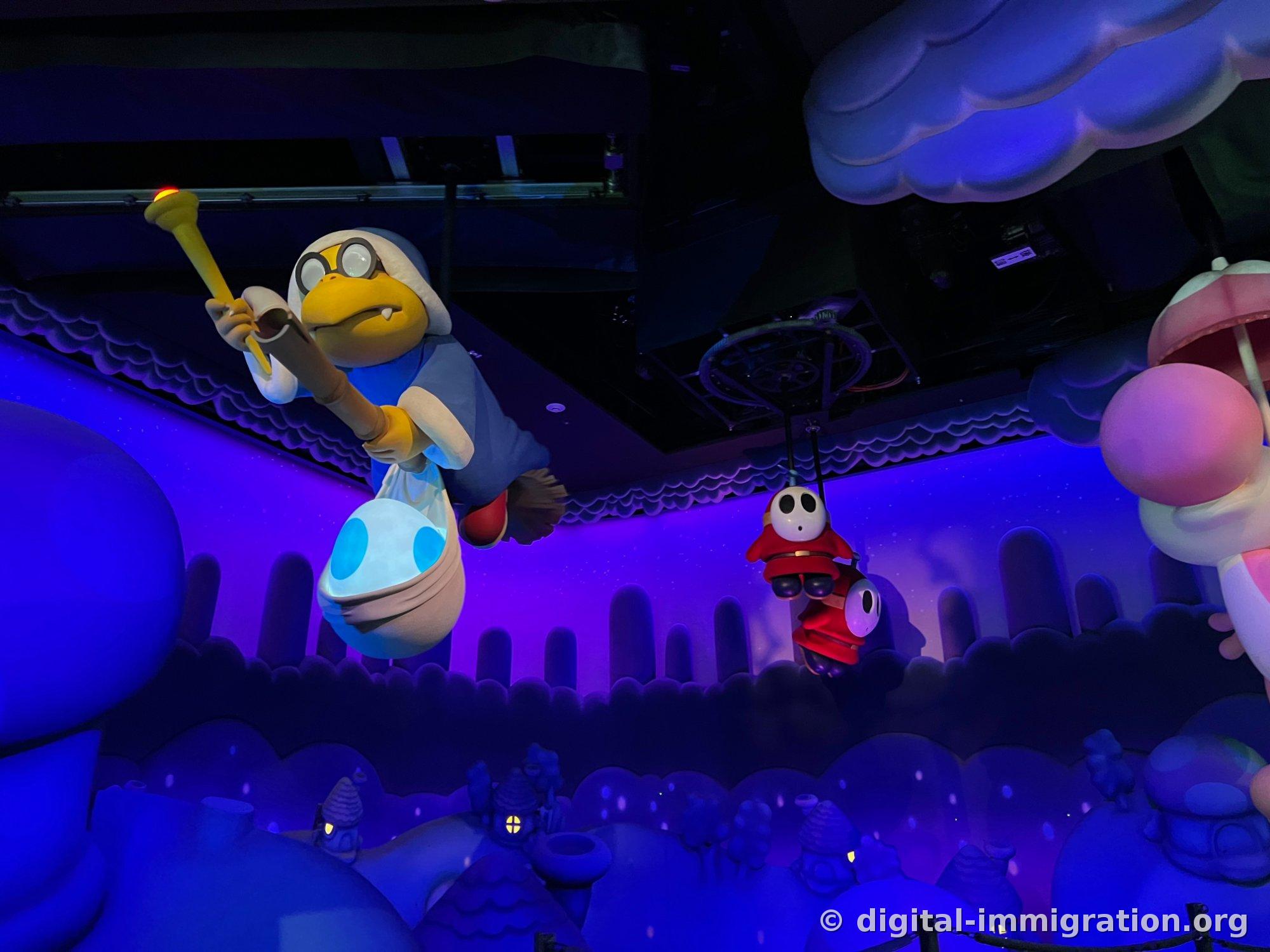 In a purple-tinted indoor section, Kamek hangs from the ceiling holding a wand and grasping an egg sack, with two red-clothed Shy Guys hanging from ropes beside him