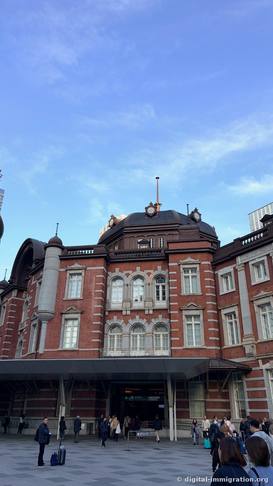 Le bâtiment en brique rouge de la Gare de Tokyo Marunouchi avec son dôme noir au-dessus de la sortie nord, le panneau JR vert, et des passants entrant et sortant