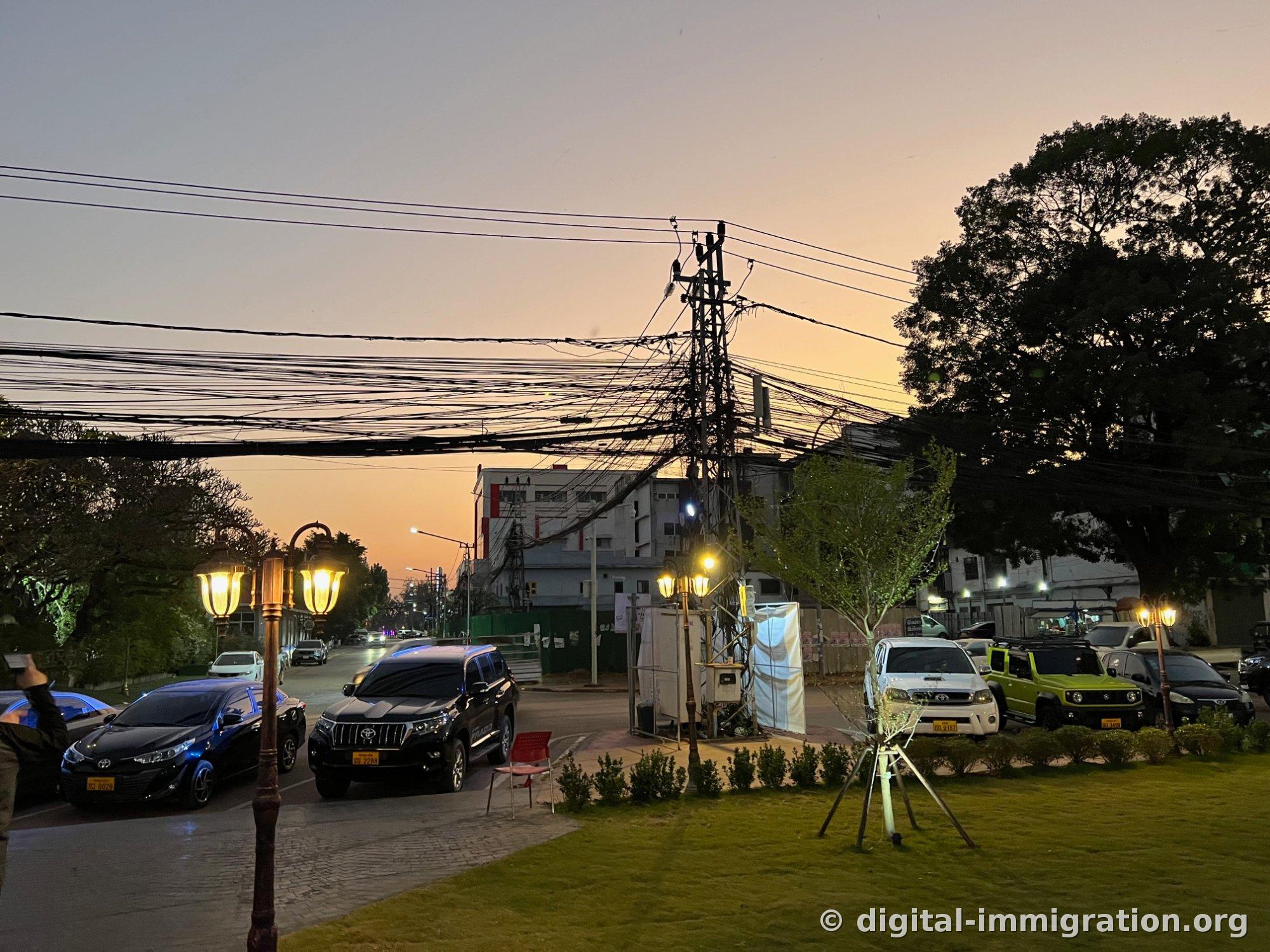 Le ciel orange du coucher de soleil avec des poteaux électriques emmêlés de câbles et des lampadaires, des SUV noirs et des camionnettes garés sur la route face à la place