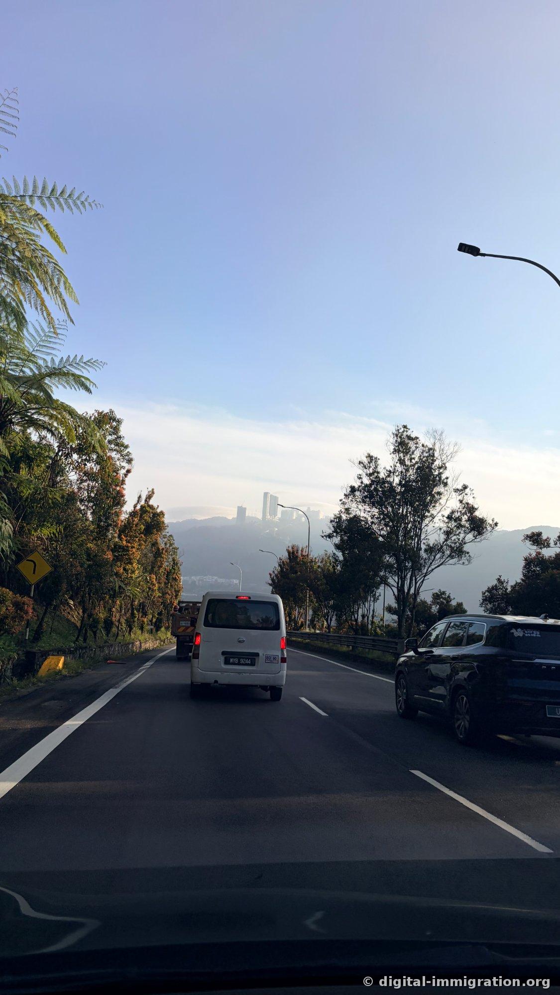 Morning mountain road view following the car ahead, with Genting's high-rise buildings visible on the ridge in the distance