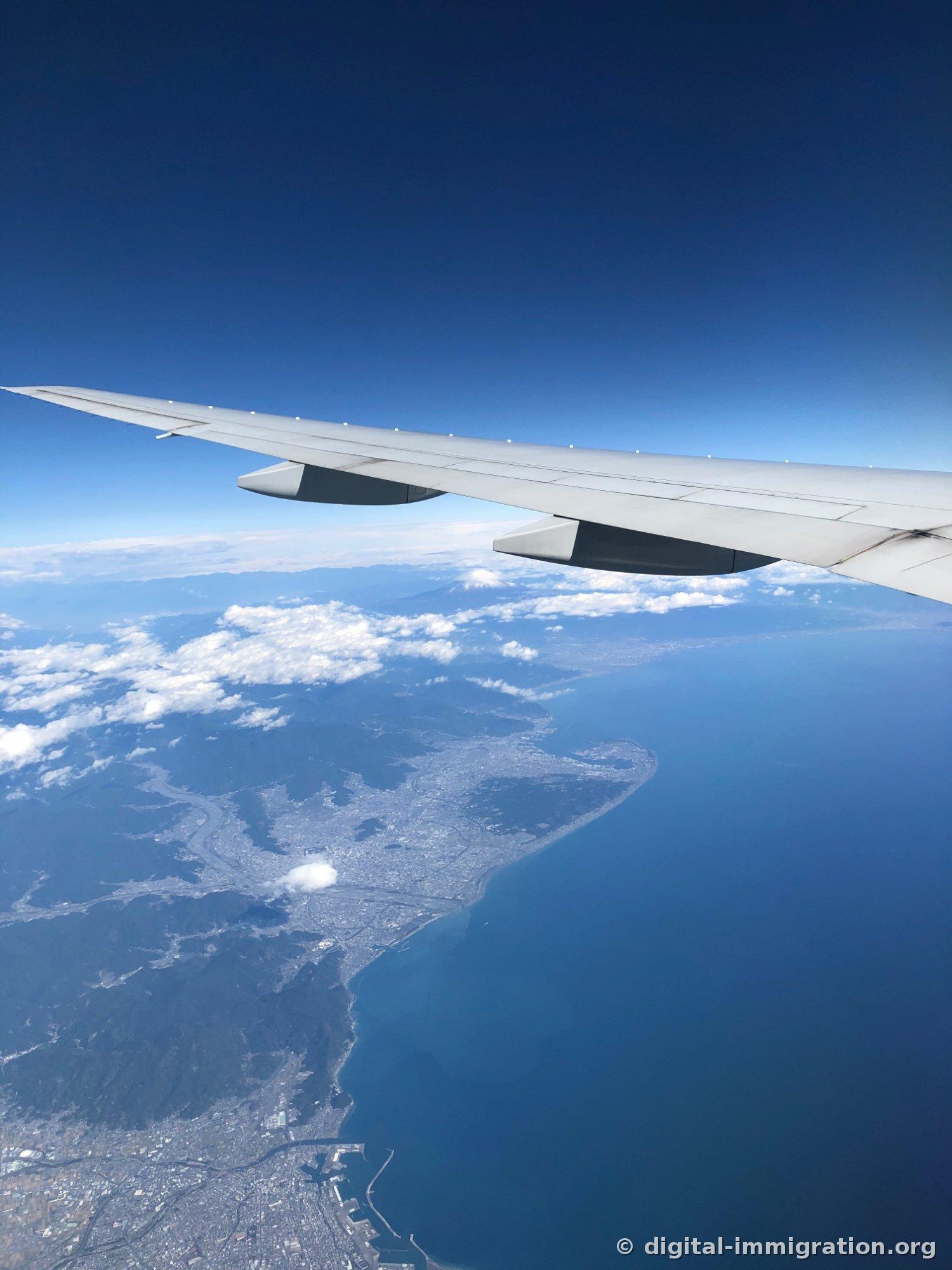 Une vue aérienne sous l'aile de l'avion montrant les eaux bleu foncé de la baie de Suruga et les côtes sinueuses, avec une zone urbaine entrelacée de rivières et le sommet du Mont-Fuji s'élevant faiblement à l'horizon au loin