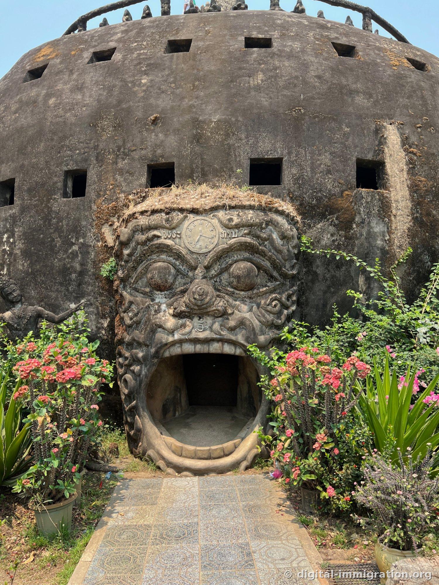 Un visage de démon géant attaché à l'avant d'un bâtiment circulaire en béton, avec la bouche béante comme passage et des pots de fleurs rouges de chaque côté