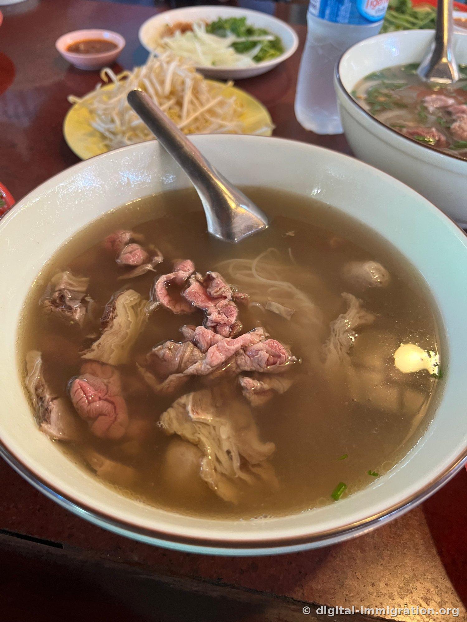 A large bowl with clear brown broth containing raw beef slices still showing a red tinge, tendons, meatballs, and rice noodles, with a bean sprout plate placed beside it