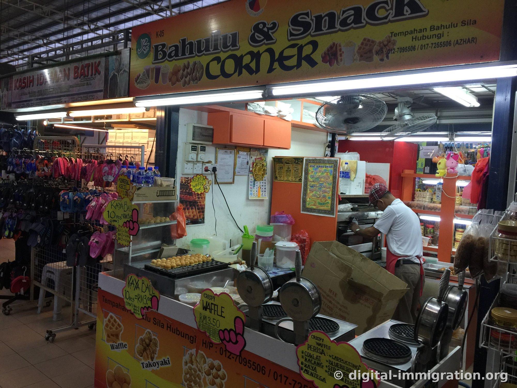Étal de pâtisseries dans le marché avec un panneau jaune « Bahulu & Snack Corner » suspendu, une machine à gaufres et une plaque pour les kaya balls en avant, avec un homme portant un foulard rouge en train de cuisiner