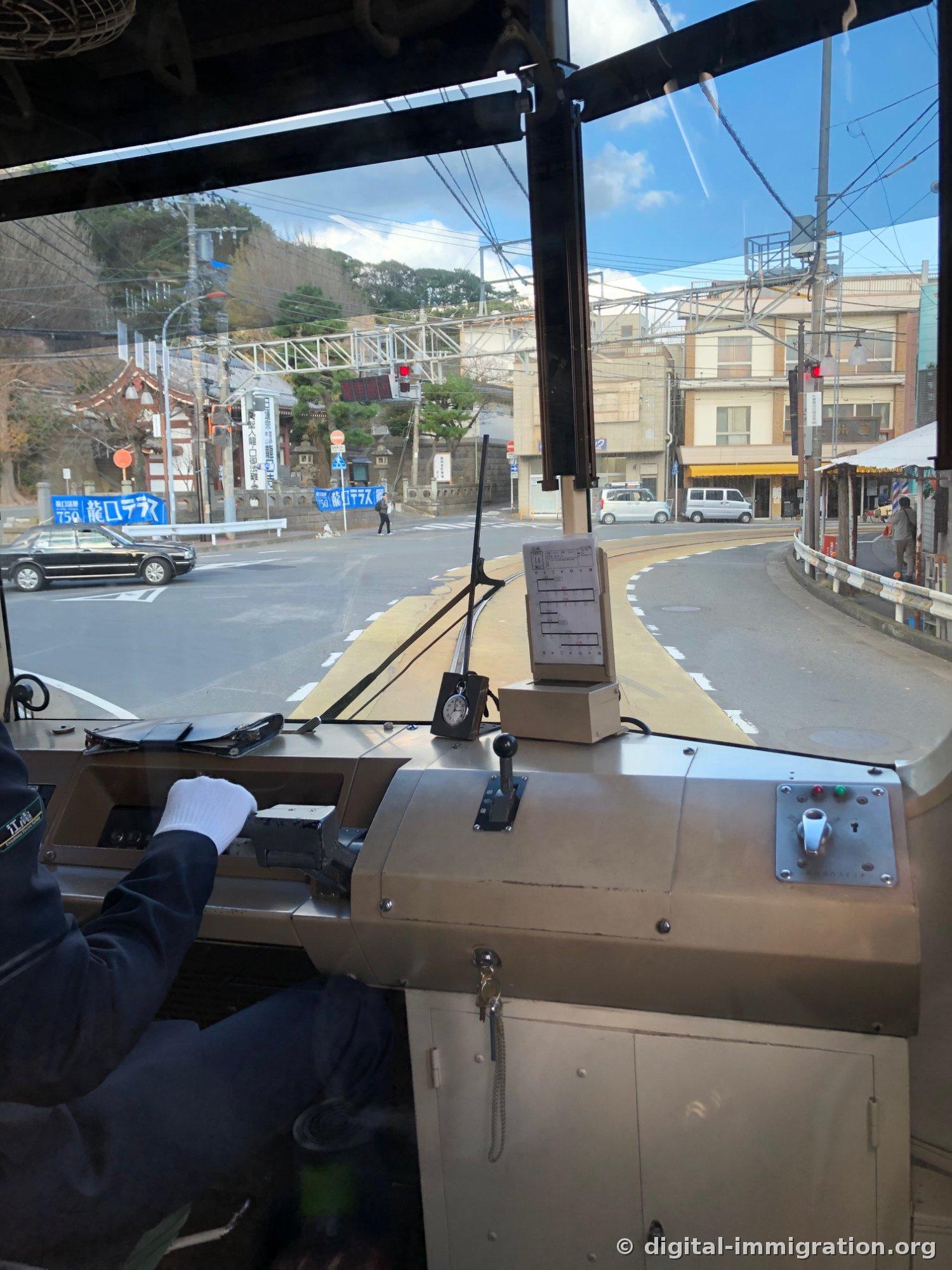 Le conducteur de l'Enoden aux mains gantées de blanc tenant le contrôleur de traction, avec l'entrée du temple Ryukoji et un panneau bleu en vue, et les rails de surface sur la route