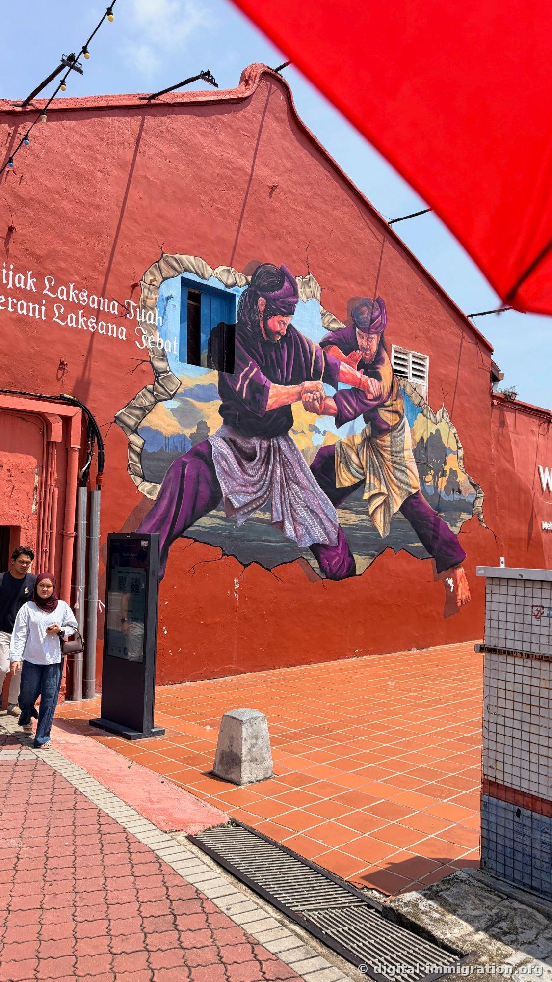 A large mural painted on a red shophouse wall depicting two figures in traditional Malay clothing in combat. Text reading 'Laksana Tuah' and 'Laksana Jebat' appears on the left. Two pedestrians pass below