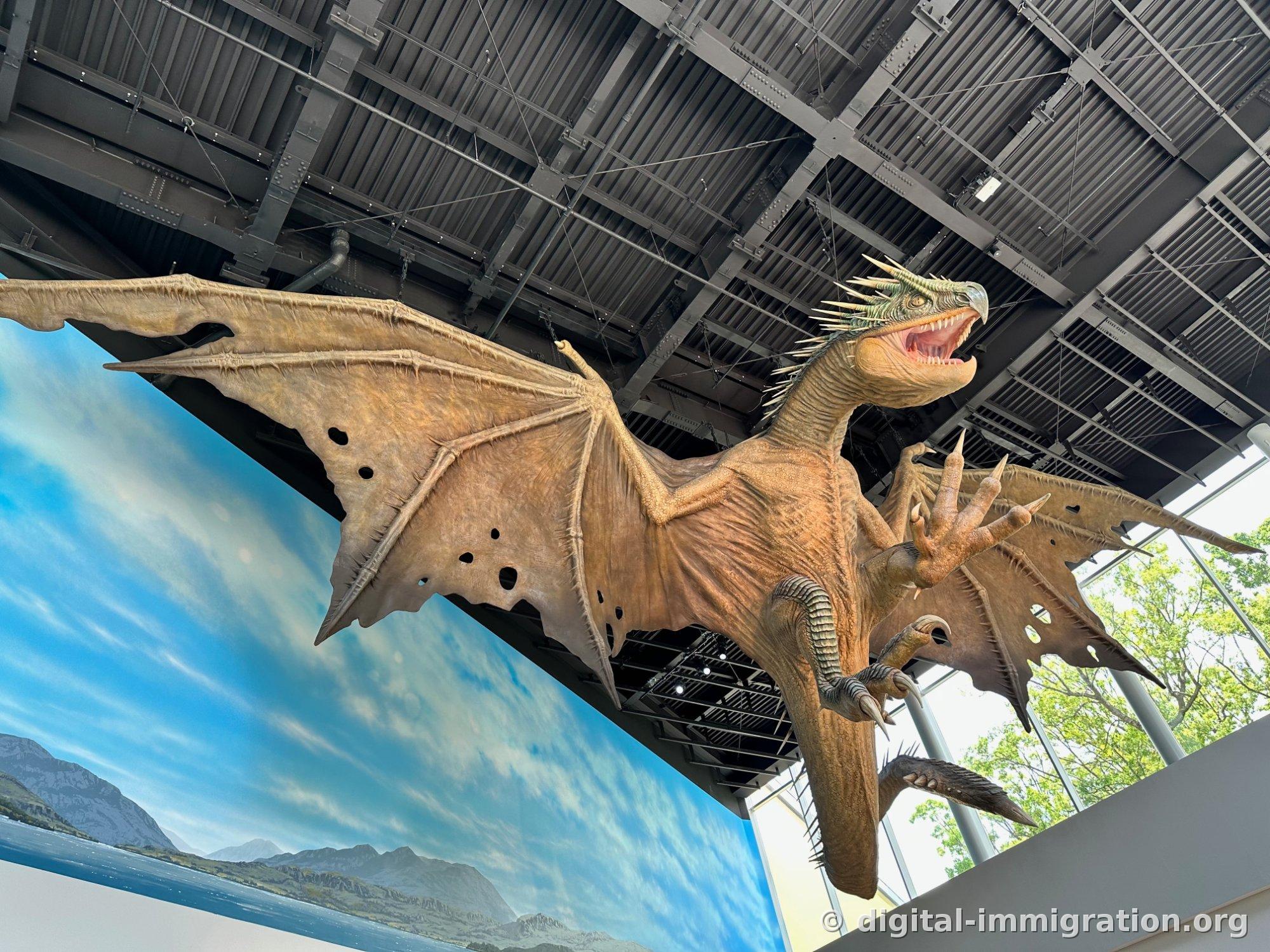 A brown dragon sculpture suspended by wire from the steel ceiling, with a painted mural of a lake and mountains behind it