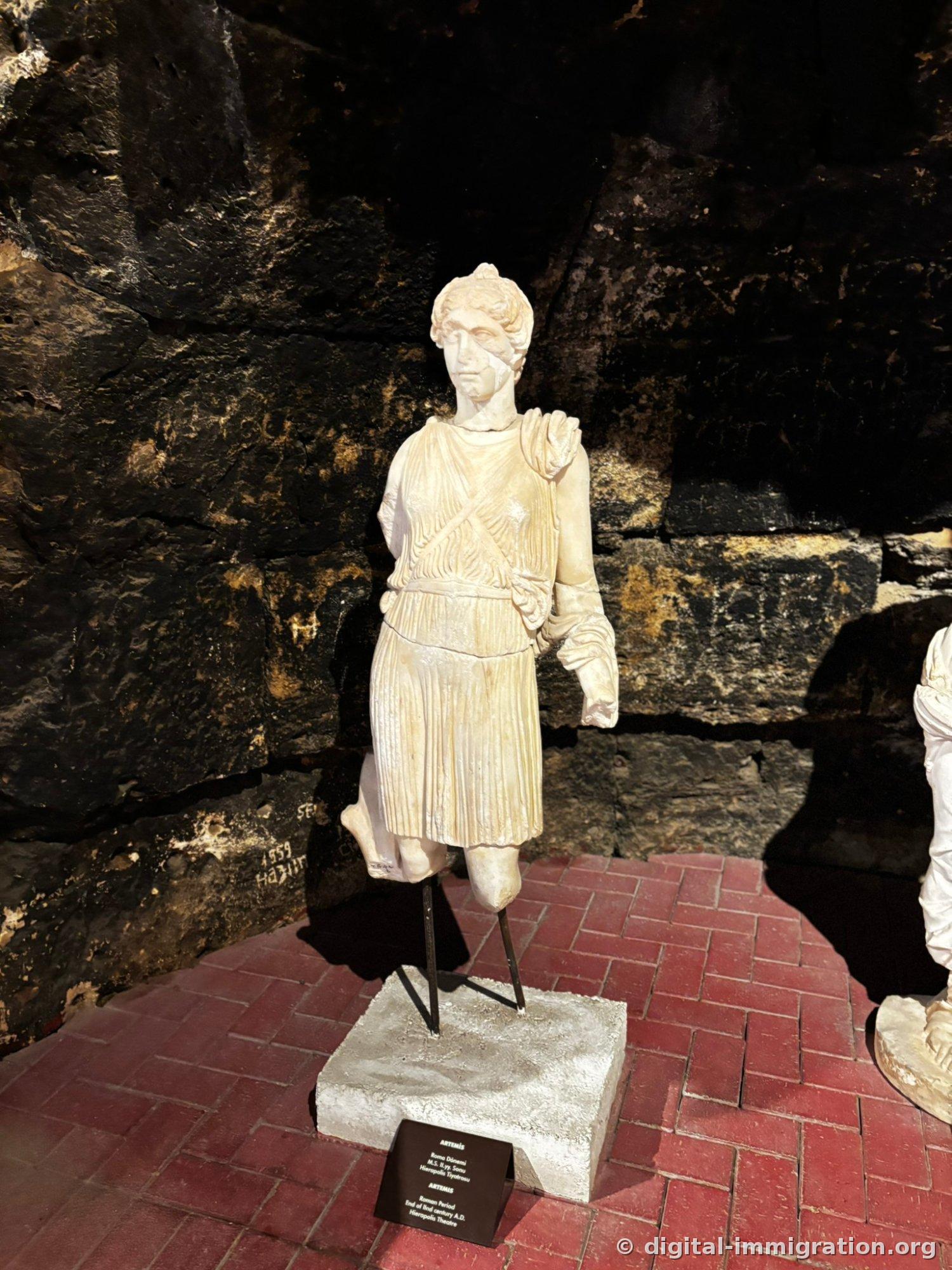 Statue of Artemis in short pleated chiton with diagonal baldric strap across her chest, mounted on metal support before a dark stone wall