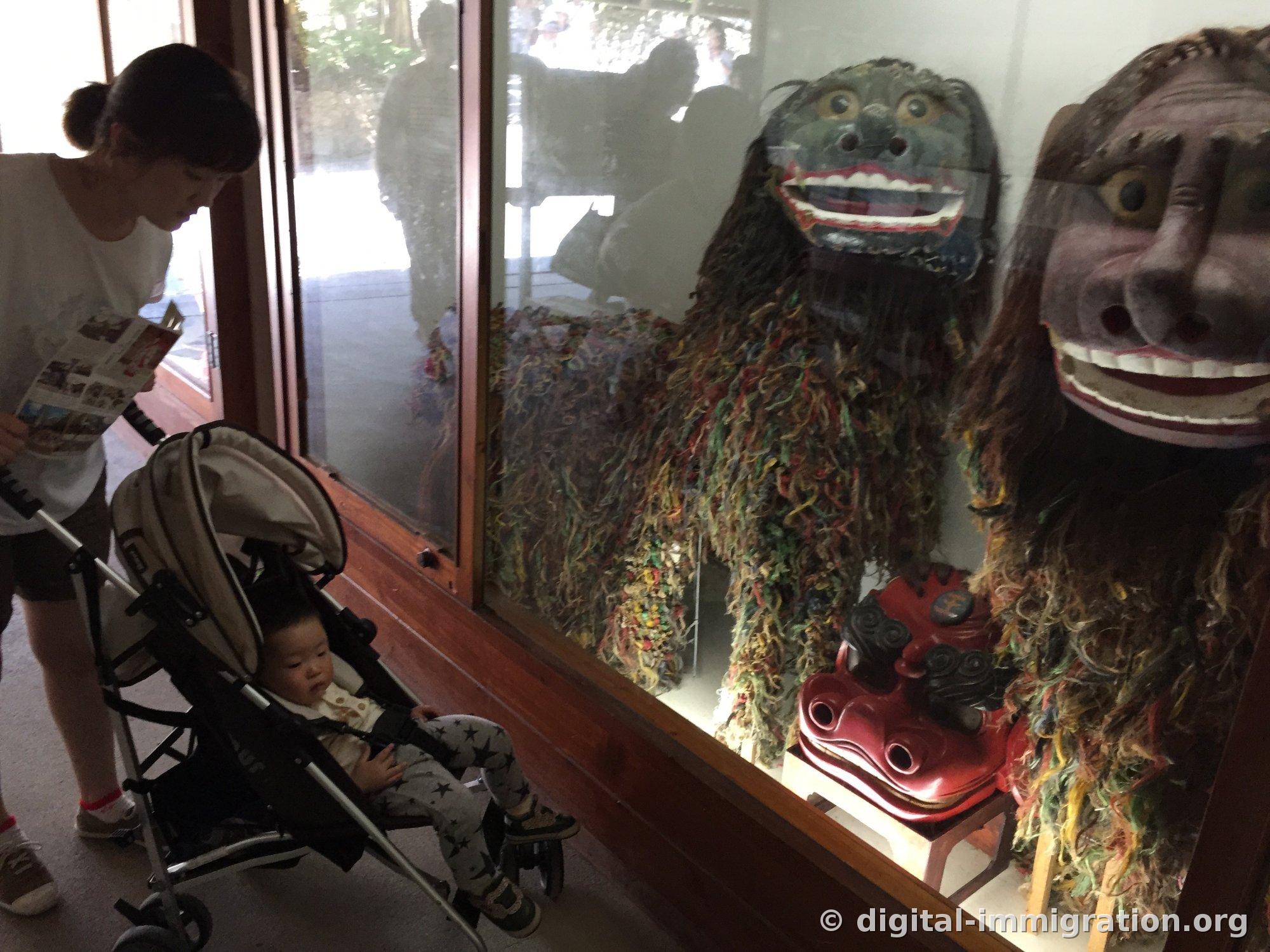 À l'intérieur d'une vitrine de verre : des masques de lion traditionnel d'Okinawa et un grand masque de visage, avec un corps fait de fils colorés entrelacés. Devant la vitrine, un visiteur regarde avec une poussette stationnée