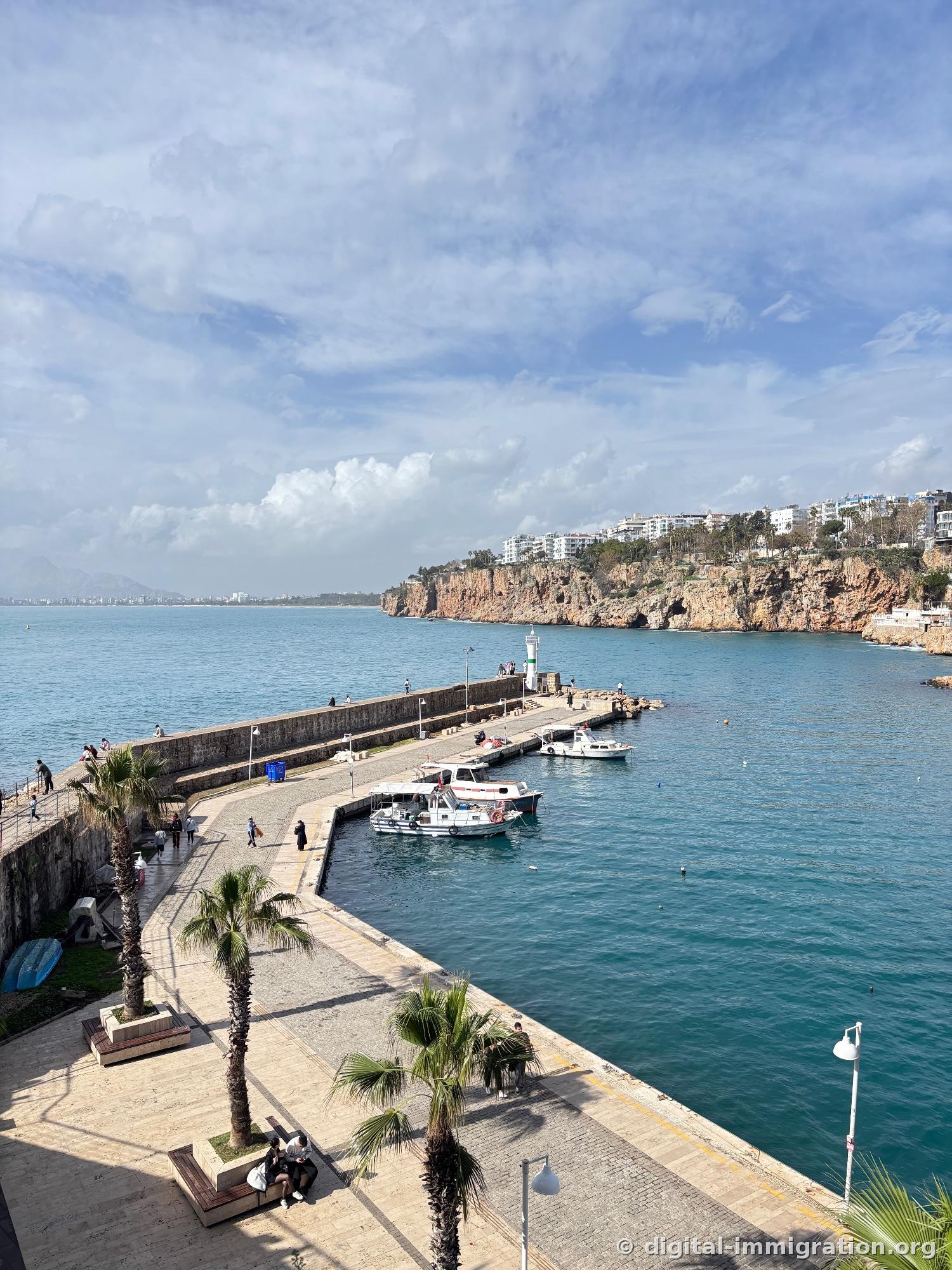 Palm-lined harbor promenade and breakwater with small lighthouse at the end, red cliffs and white buildings extending along the coast on the right