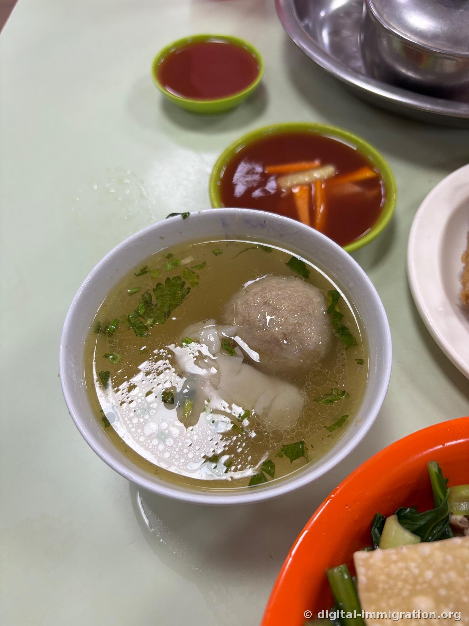 Petit bol de bouillon clair contenant une boulette de bœuf grise et une boulette de pâte bouillie avec feuilles de céleri, coupelle de sauce sambal rouge et plat de carottes marinées à côté