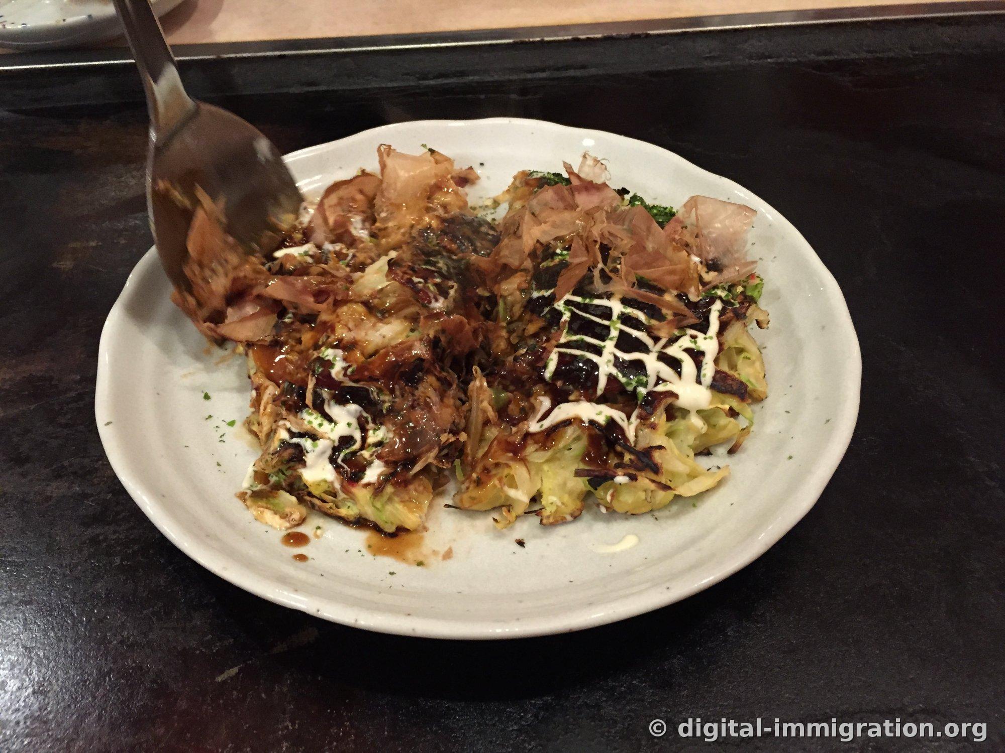 Okonomiyaki on a white plate on a black teppan grill. Brown sauce and mayonnaise drizzled in a crosshatch pattern, topped with aonori and bonito flakes, with a teppan spatula beside it