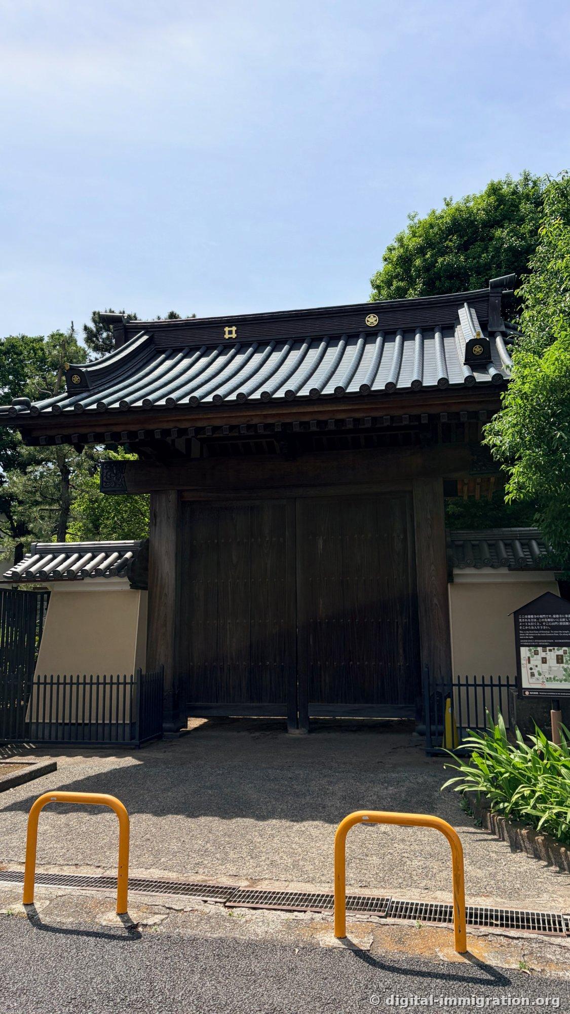 The east gate of Gotokuji with a black tile roof and thick wooden doors, with two yellow bollards in front