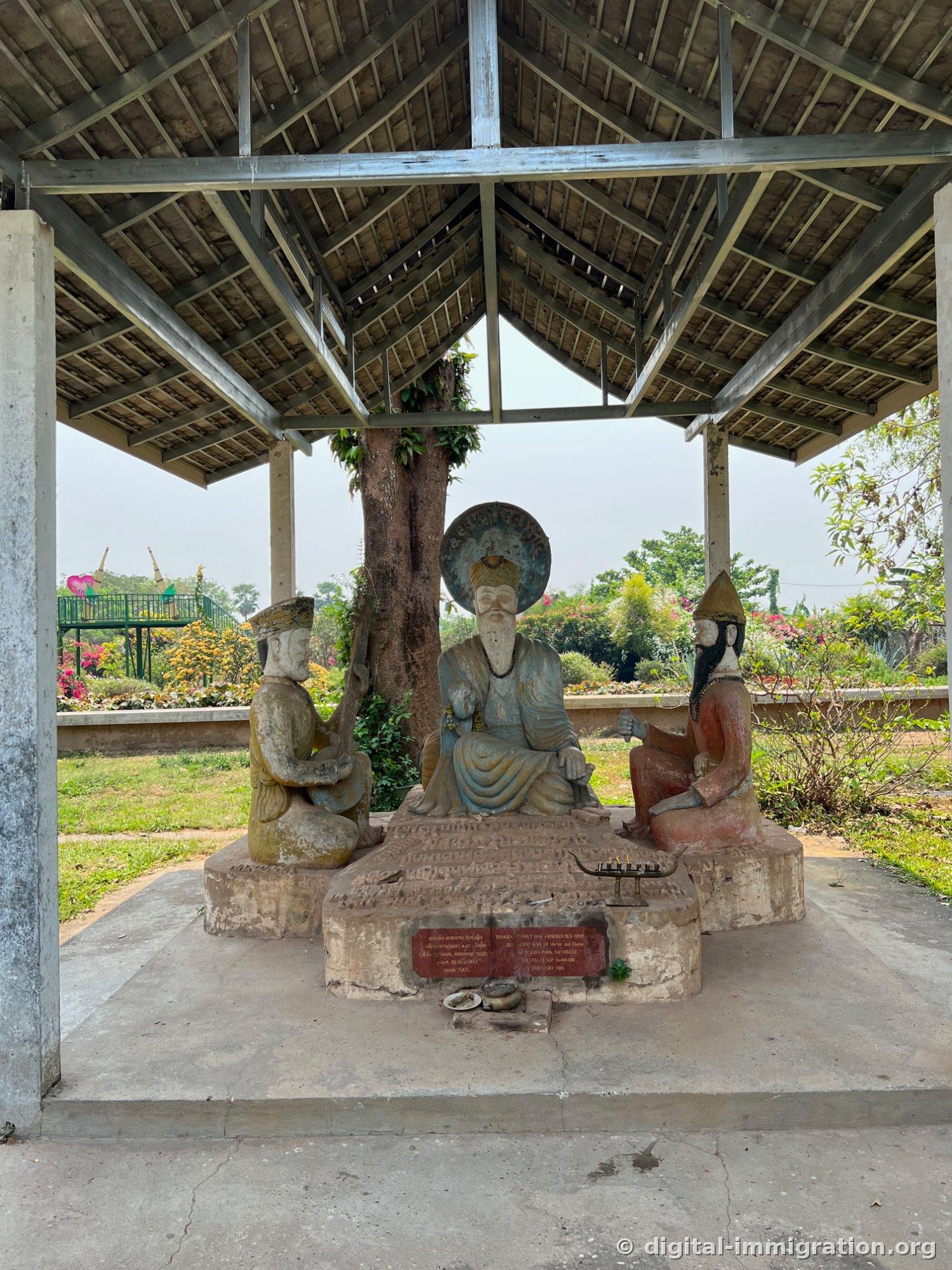 Sous un appentis à toit de tôle, une statue de figure barbe longue assise au centre et deux figures agenouillées de chaque côté, avec une plaque de donation rouge devant