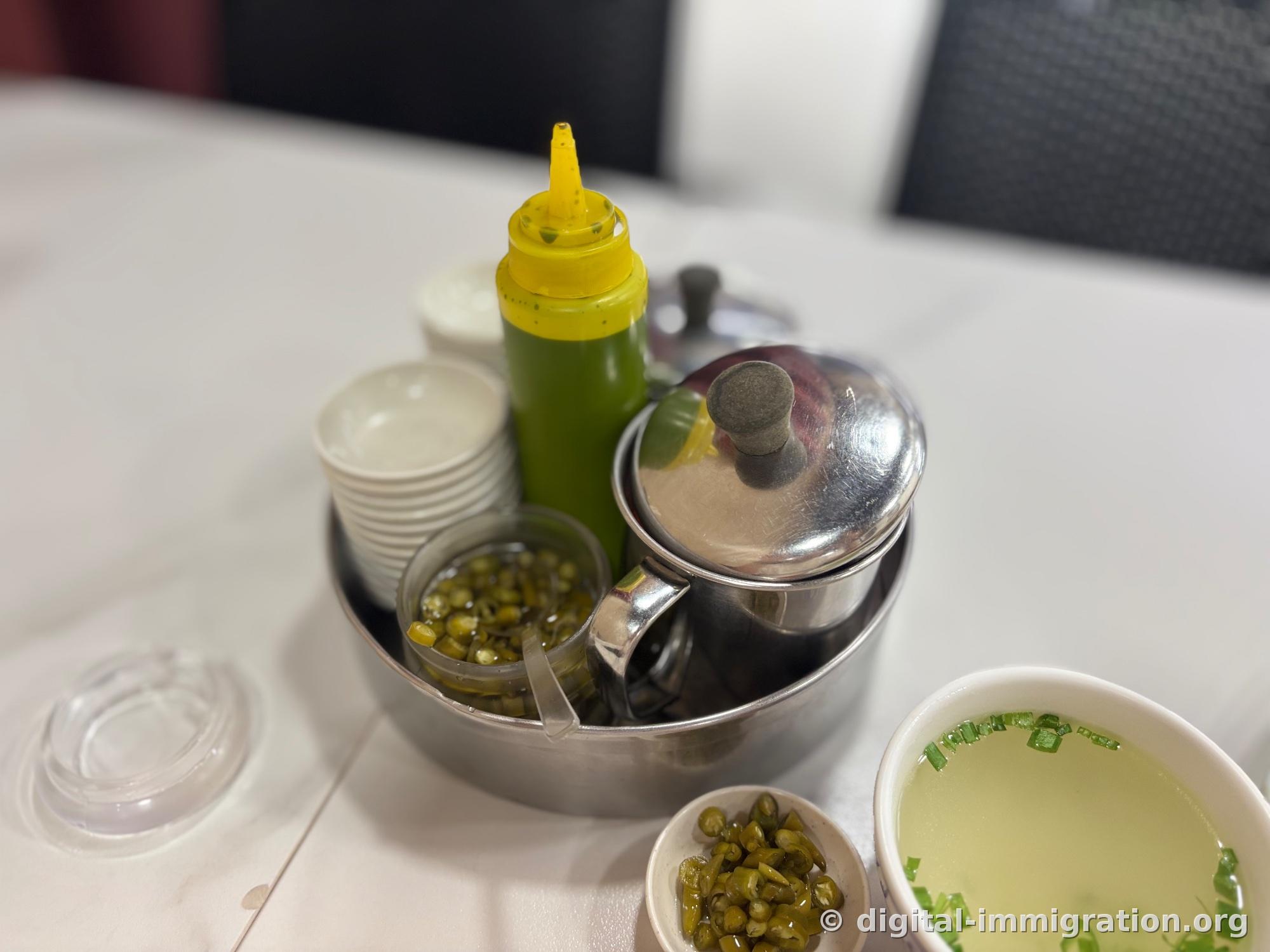 Stainless steel tray on the table with a soy sauce pot, green chili sauce bottle with yellow cap, glass cup of pickled chilies, and stacked white bowls