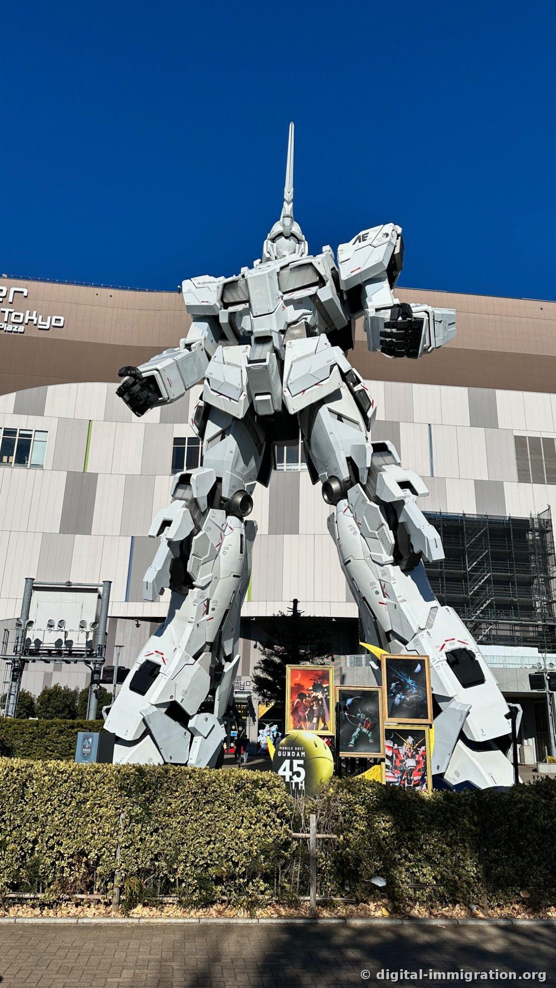 A full-size white Unicorn Gundam standing frontally in front of the DiverCity Tokyo Plaza building, with a single horn on its head in closed Unicorn mode