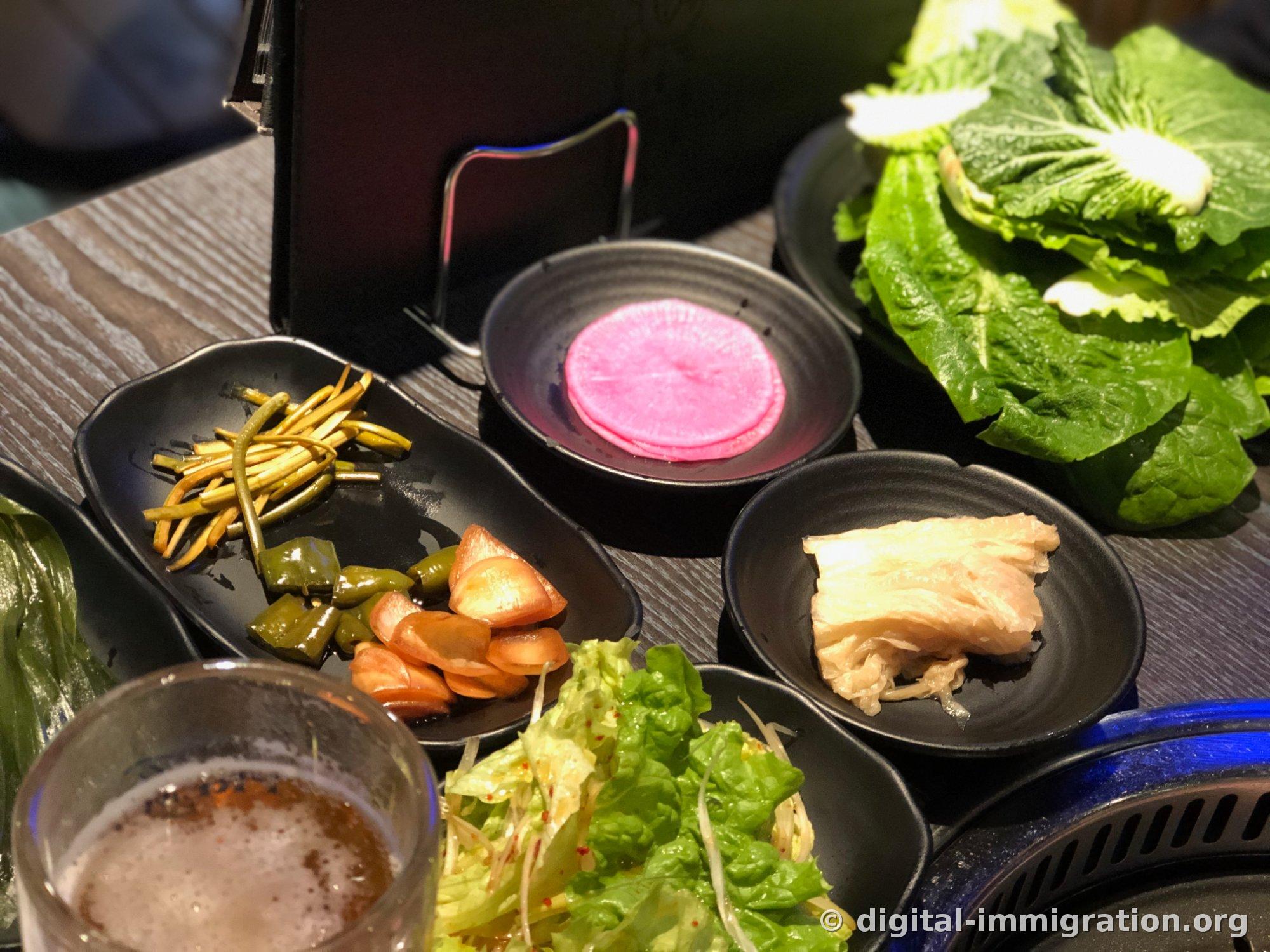 黒いお皿に盛られたニンニクの芽の漬物、きゅうりの漬物、プチトマトの漬物とピンク色のサムとり、白キムチ、サンチュと青じそ、ネギの浅漬けが置かれた焼肉屋のテーブル