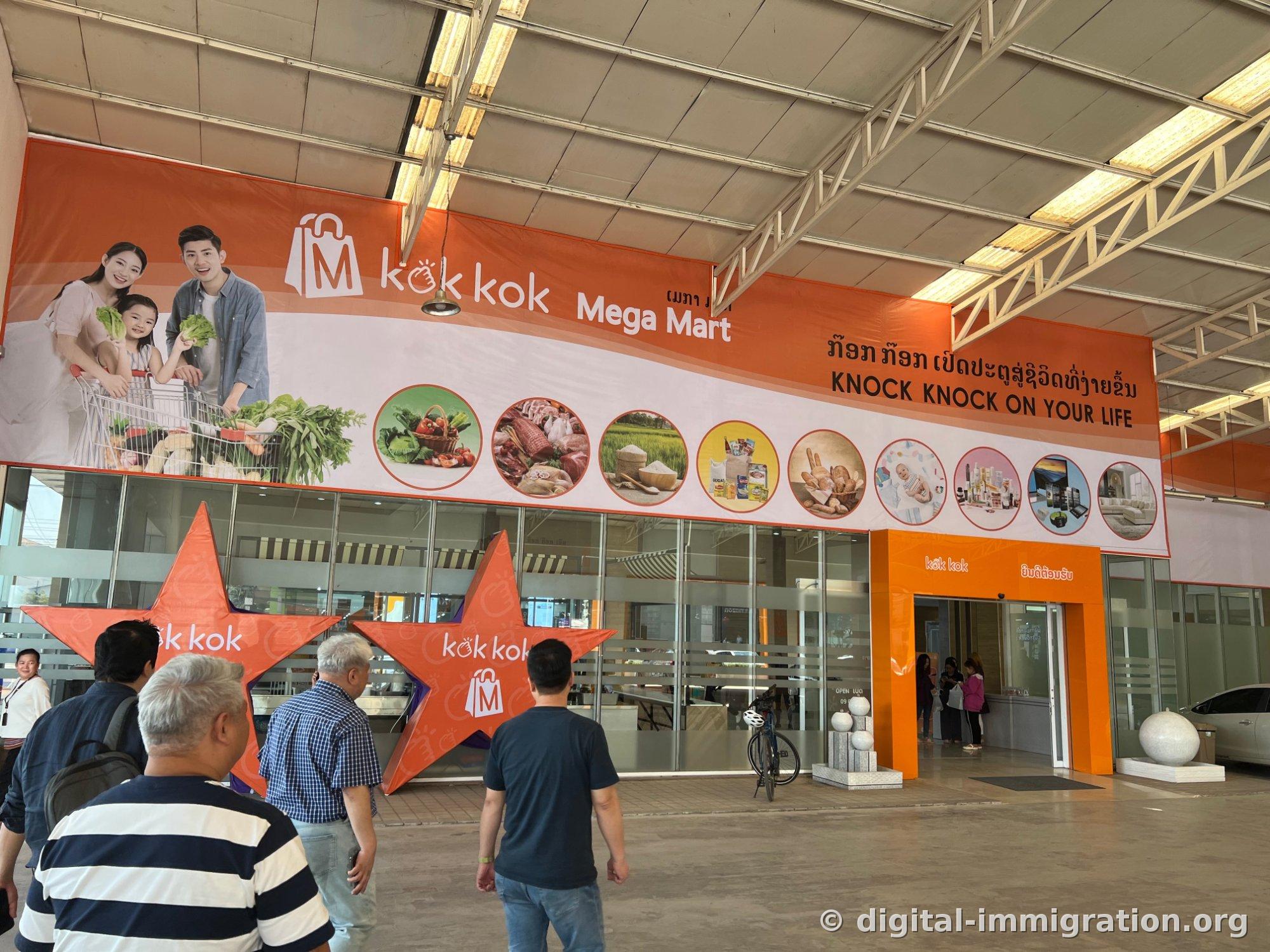 지붕 덮인 진입로 위에 걸린 대형 주황색 kok kok Mega Mart 현수막과 별 모양 조형물, 자동문 입구로 사람들이 걸어 들어가는 모습