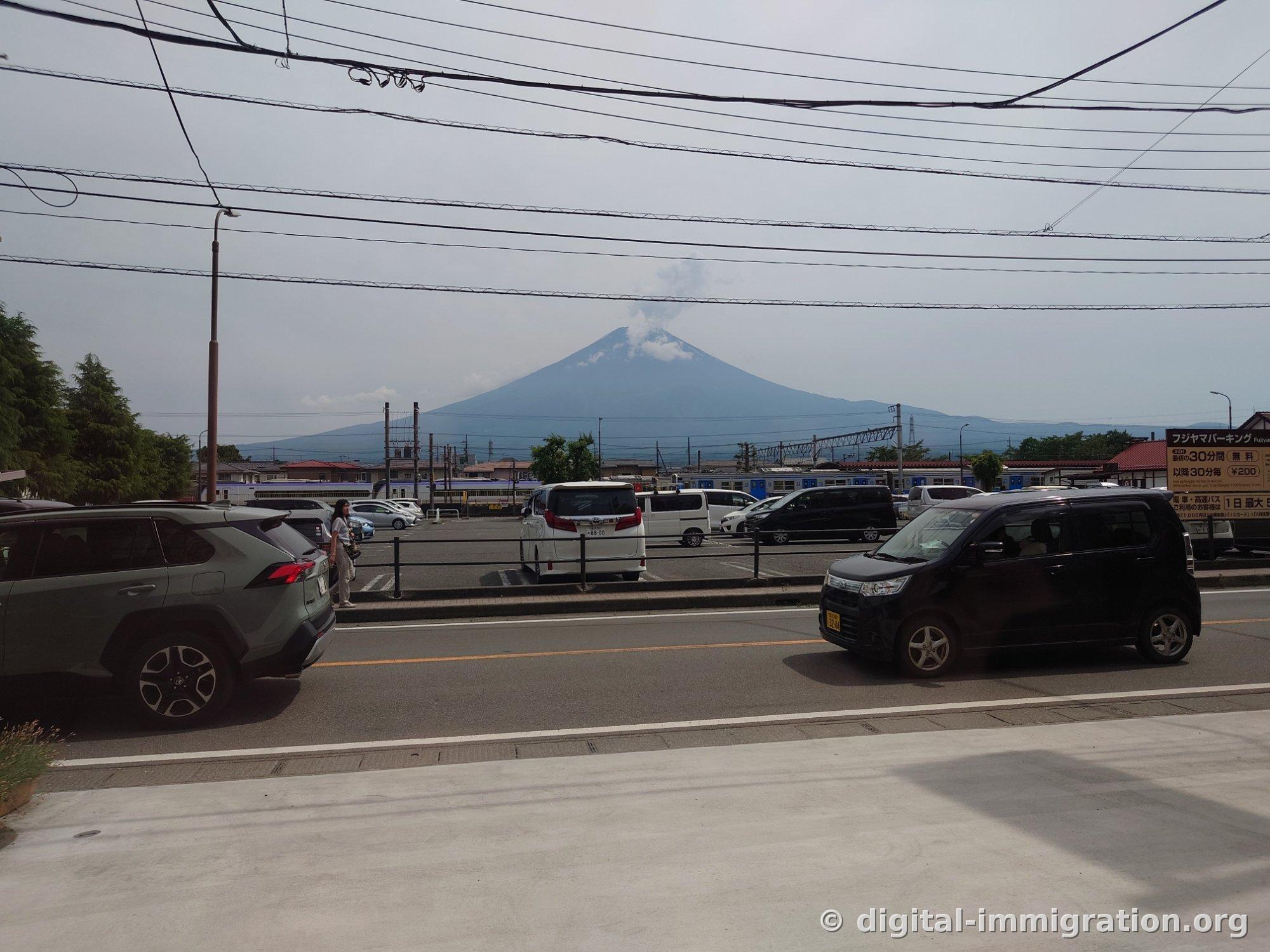 道路の向こう側の富士山パーキング駐車場と料金案内看板、その向こうに富士山の全景