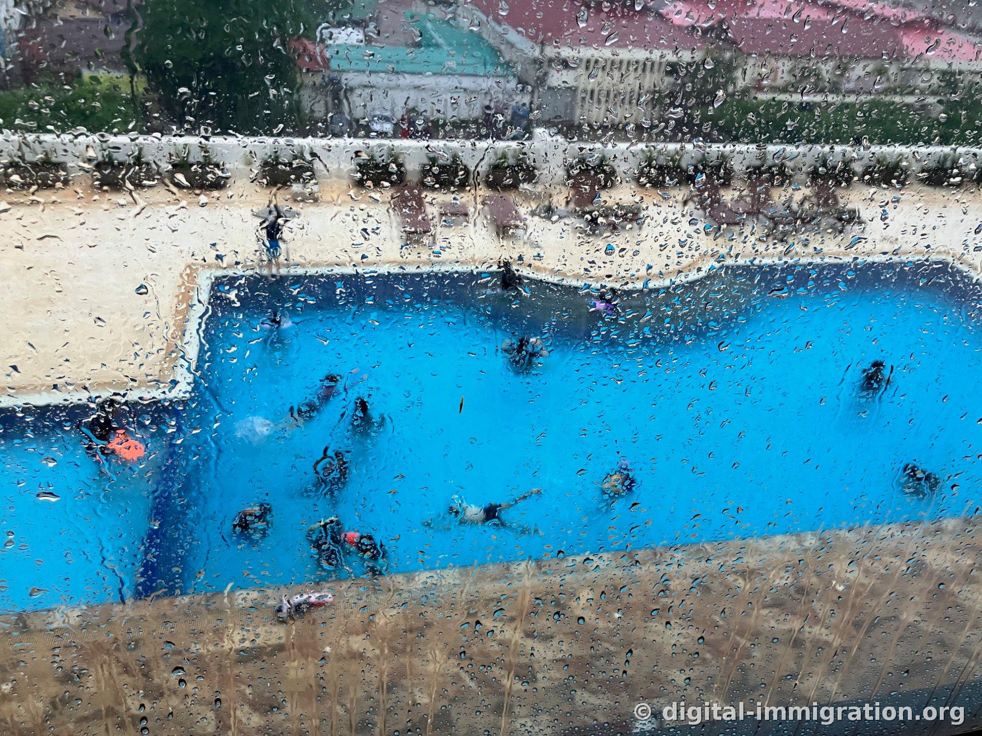 Vue de la piscine extérieure bleue avec nageurs à travers une fenêtre couverte de gouttes de pluie