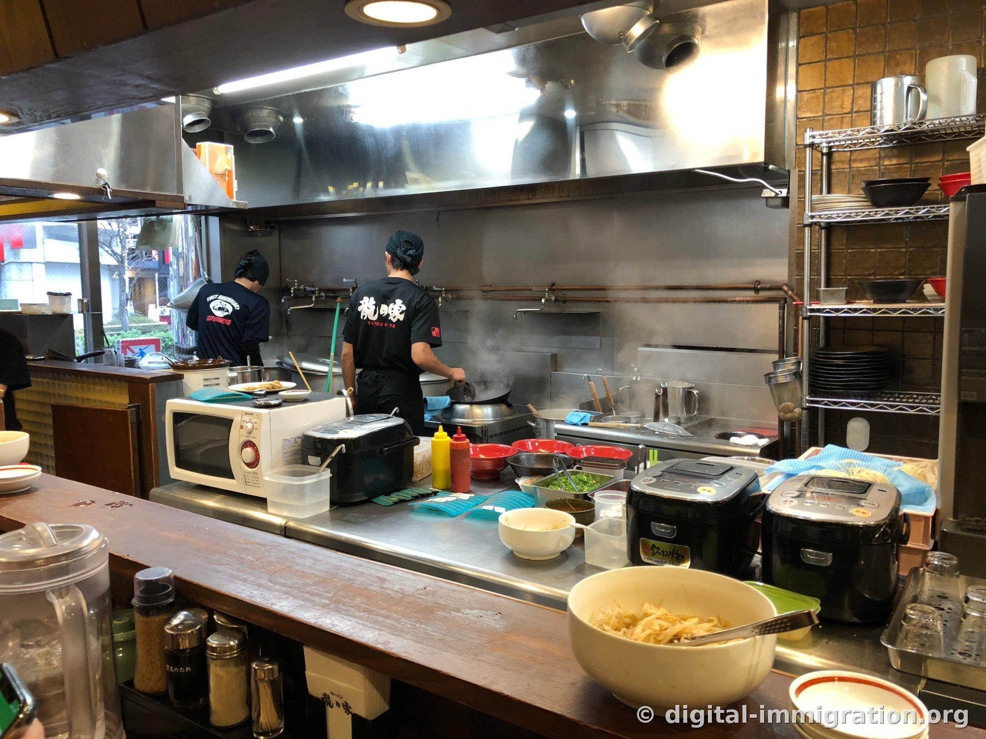 La cuisine ouverte vue du comptoir. Deux employés vêtus de t-shirts noirs avec le logo 龍の家 travaillent devant des foyers fumants