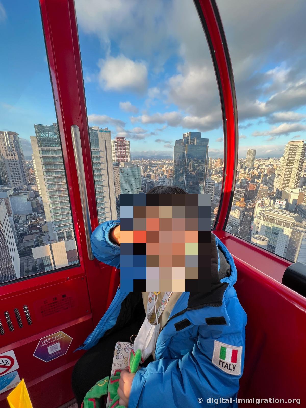 赤いカプセル内部で、青いダウンジャケットを着た子どもが、窓の外の梅田高層ビルを背景に座っている様子