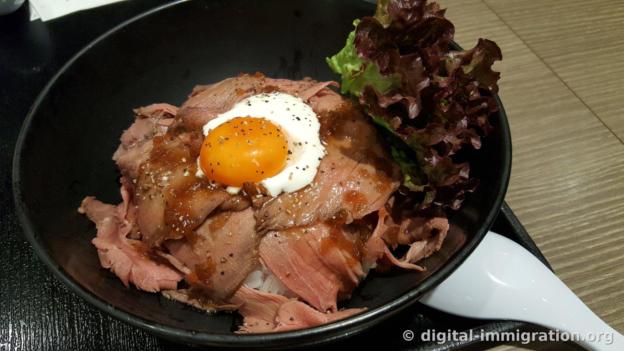 Roast beef rice bowl in a black bowl with thinly sliced pink beef, a soft-boiled egg yolk, coarse black pepper, and brown sauce on top, with red leaf lettuce on the side