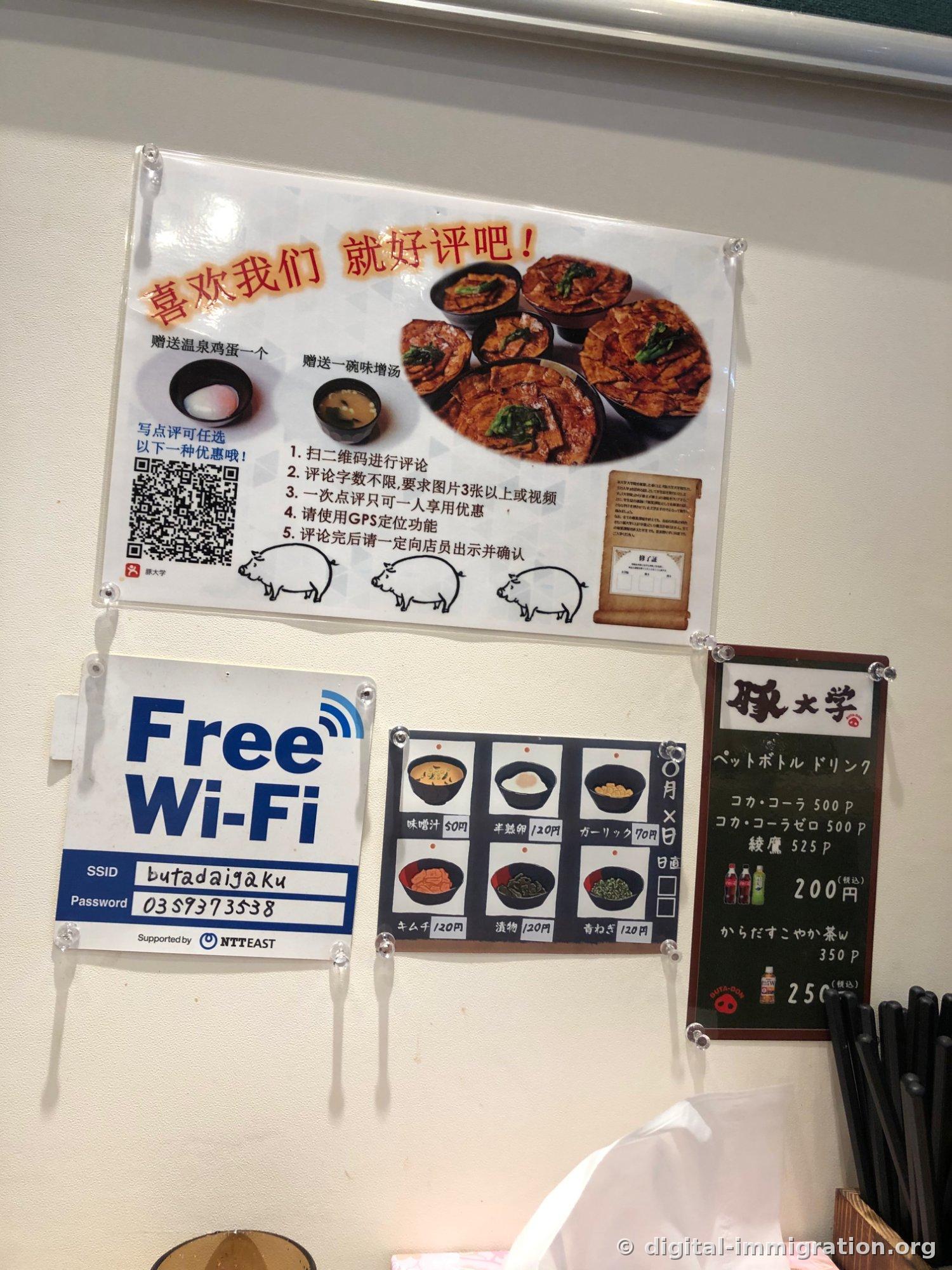 Les affichages au mur. L'affiche d'événement pour avis en chinois, l'indication du Wi-Fi gratuit SSID butadaigaku, un tableau des prix des garnitures dessiné montrant soupe miso 50 yen, œuf à moitié cuit 120 yen, ail 70 yen, et un tableau des prix des boissons en bouteille
