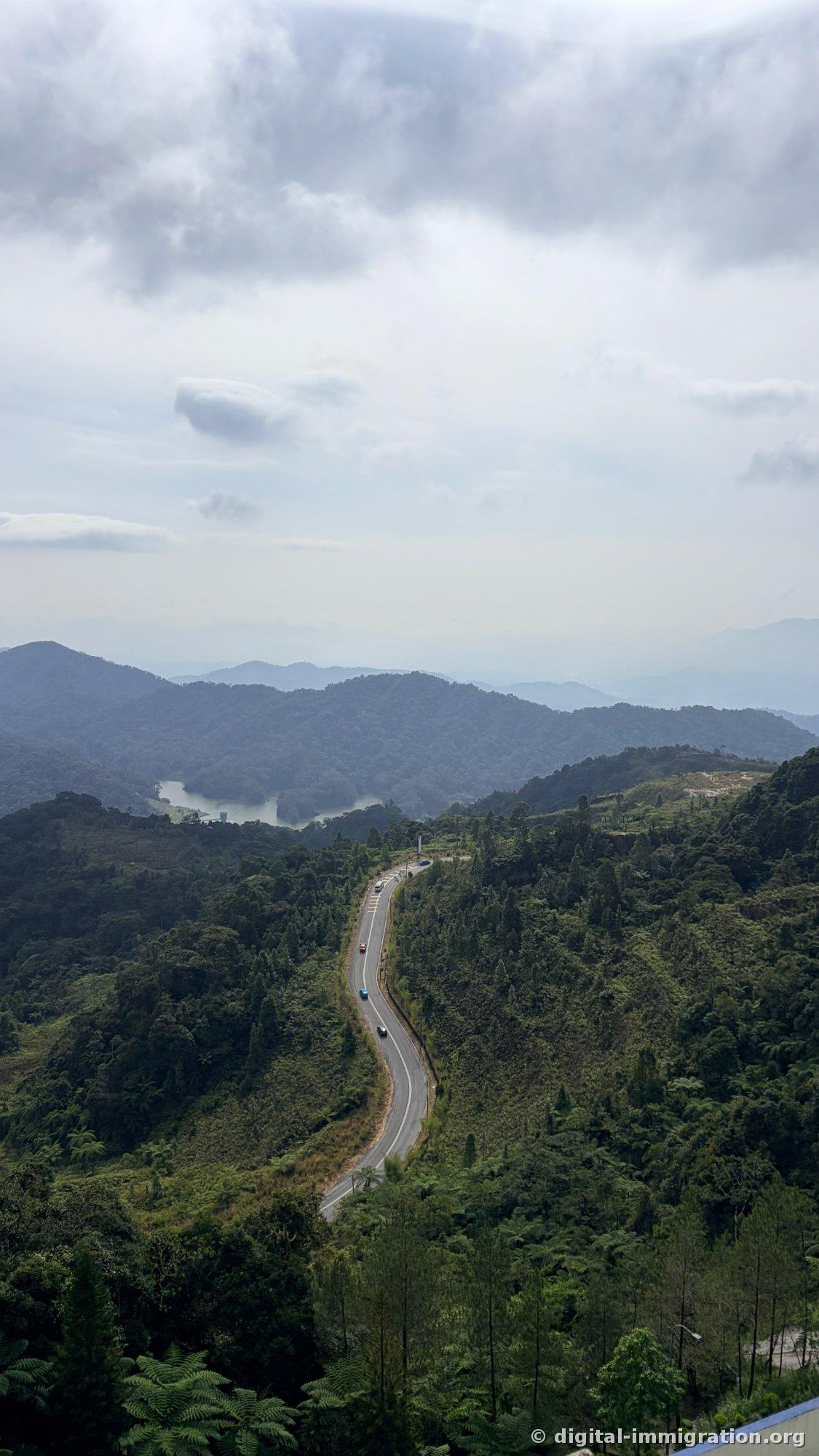 Layered mountain ridges under cloudy sky with reservoir nestled between them and S-curved mountain road