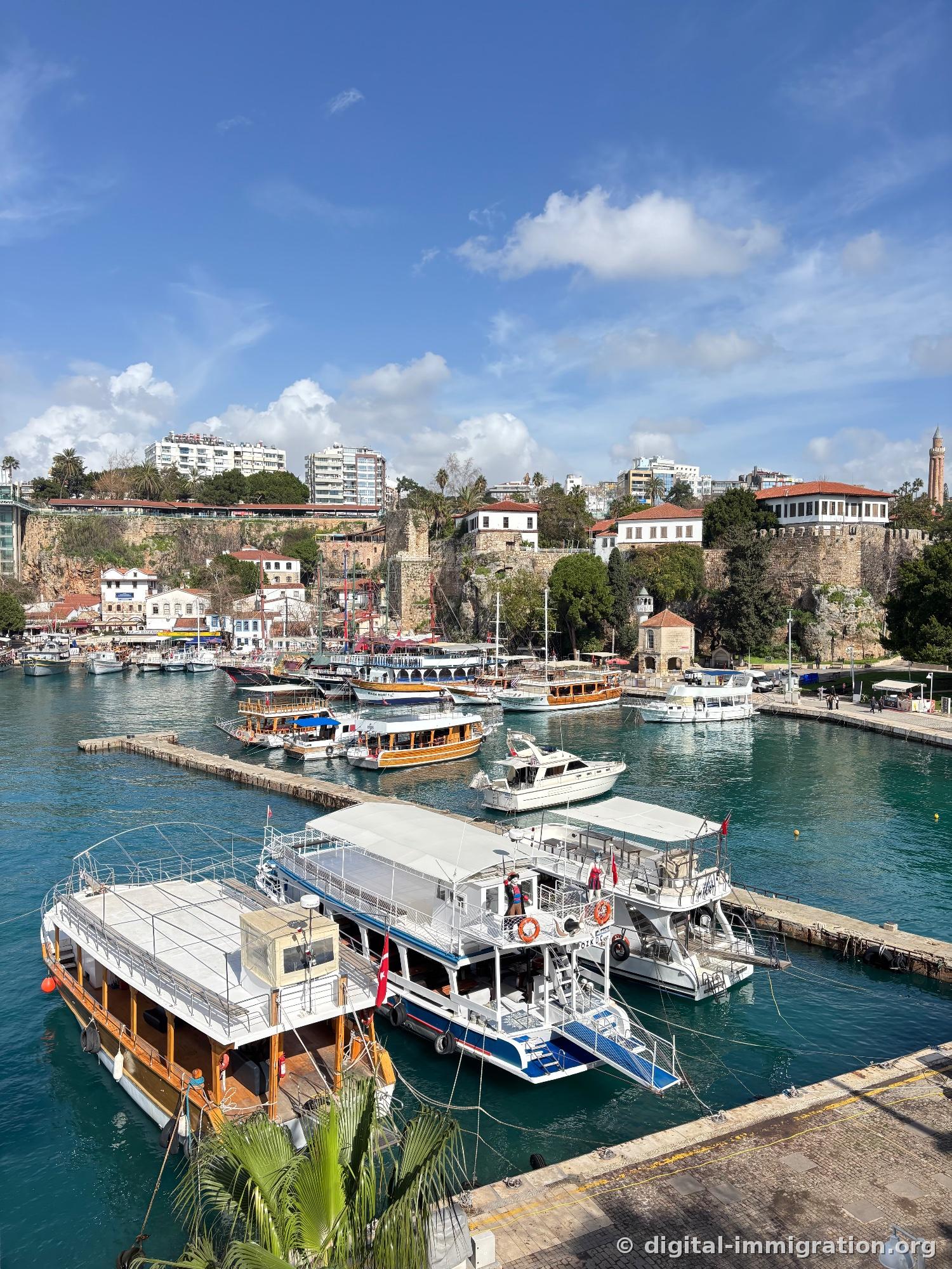 White tour boats and wooden sailboats moored in the harbor, city walls and Ottoman mansions behind, distant Yivli Minare minaret on the right