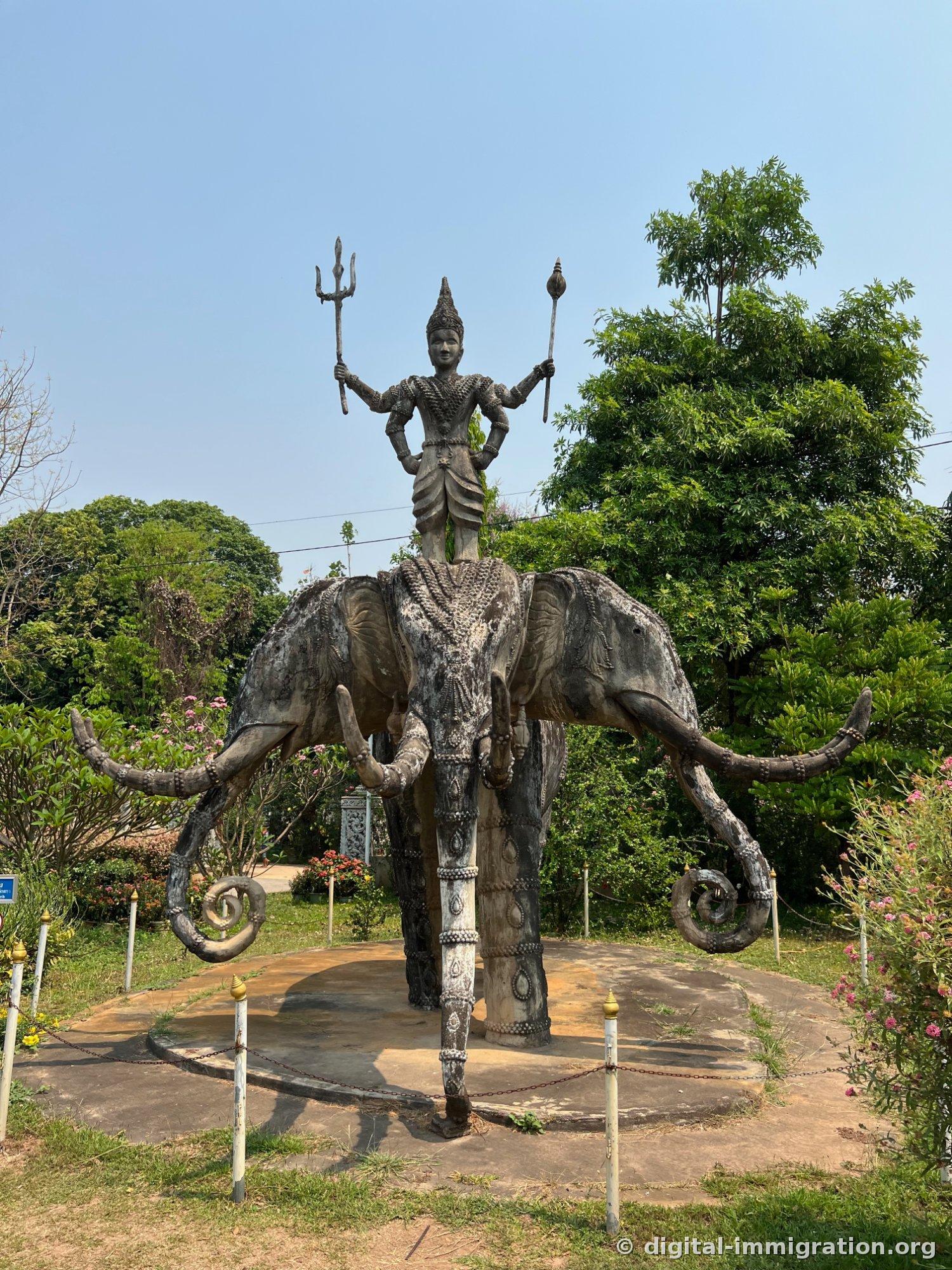 Une sculpture d'une divinité tenant un trident debout sur un éléphant à trois têtes, avec un ciel bleu en arrière-plan