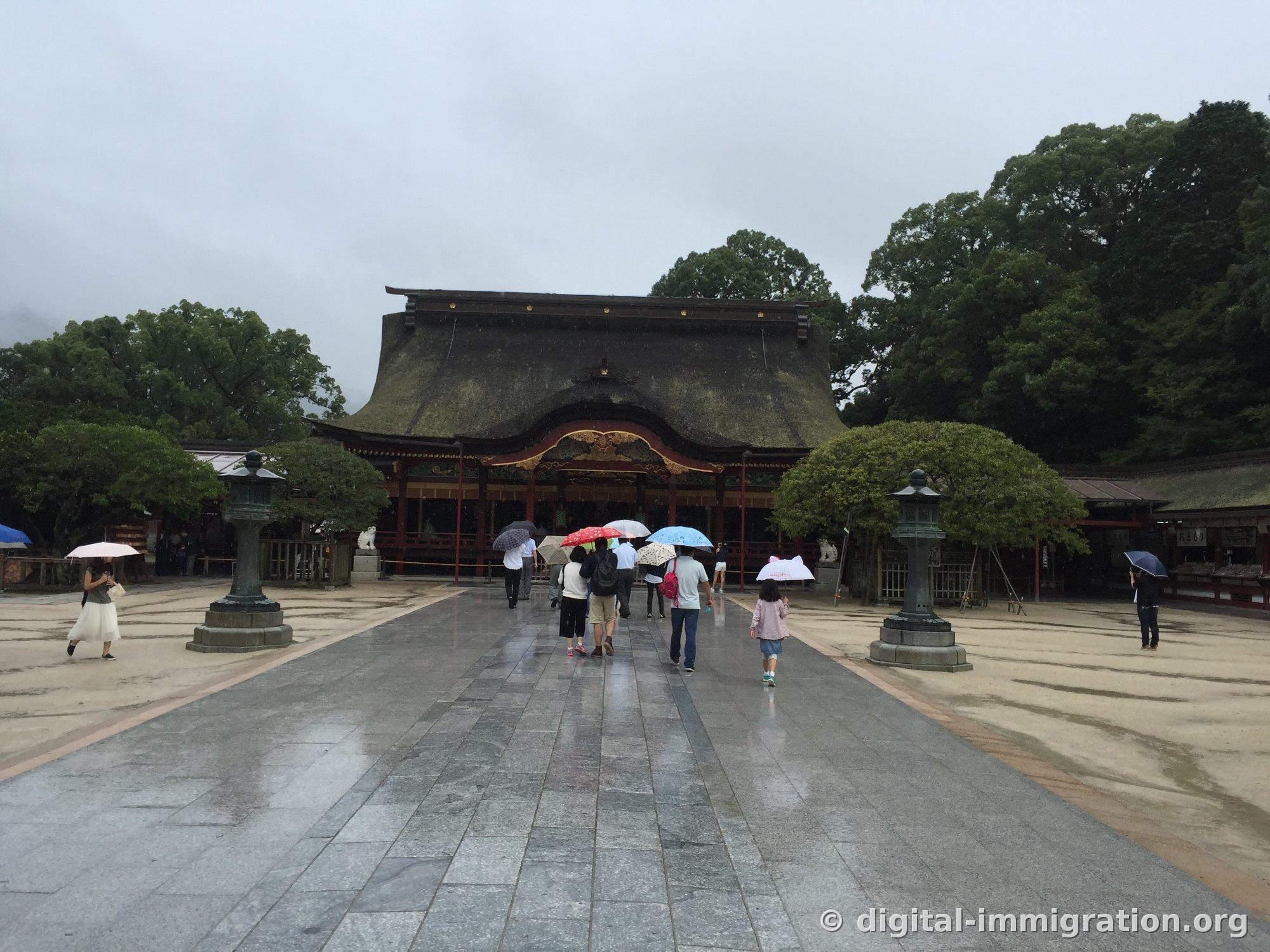 Dazaifu Tenmangu main hall visible at the end of the wet stone approach, bronze lanterns on both sides, worshippers with umbrellas