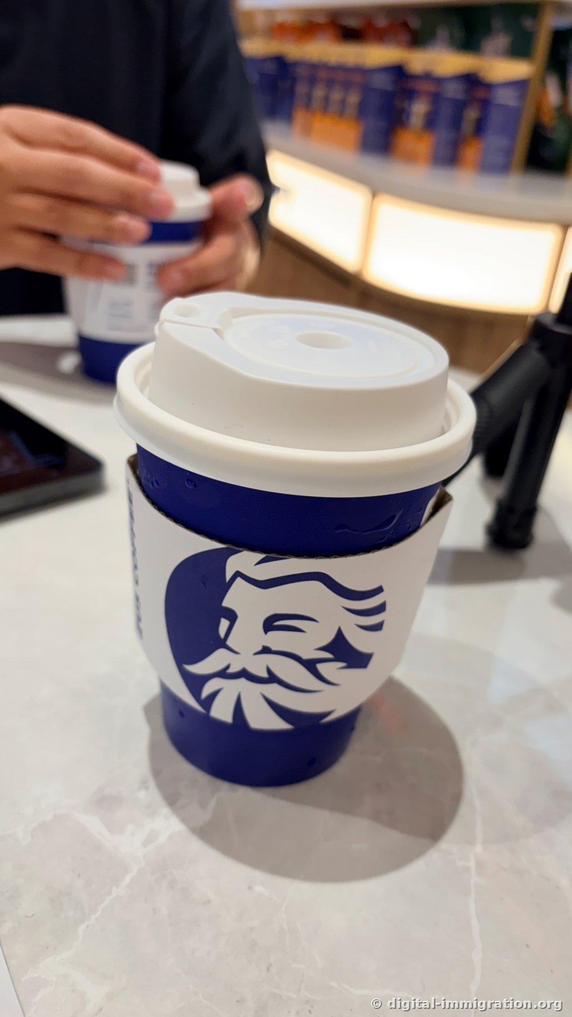 A ZUS Coffee blue cup on a marble table. The white lid and Zeus face logo sleeve are visible, and behind it someone else is opening the lid of the same cup.