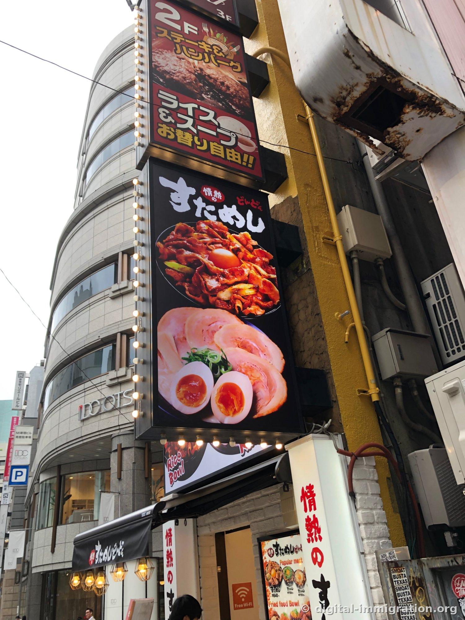 L'enseigne '情熱の すためし どんどん' accrochée verticalement sur le mur extérieur d'un bâtiment à Shinjuku, avec la publicité '2F Steak & Hamburger' en haut, et le magasin IDC OTSUKA avec son bâtiment en verre incurvé visible à l'arrière-plan à gauche