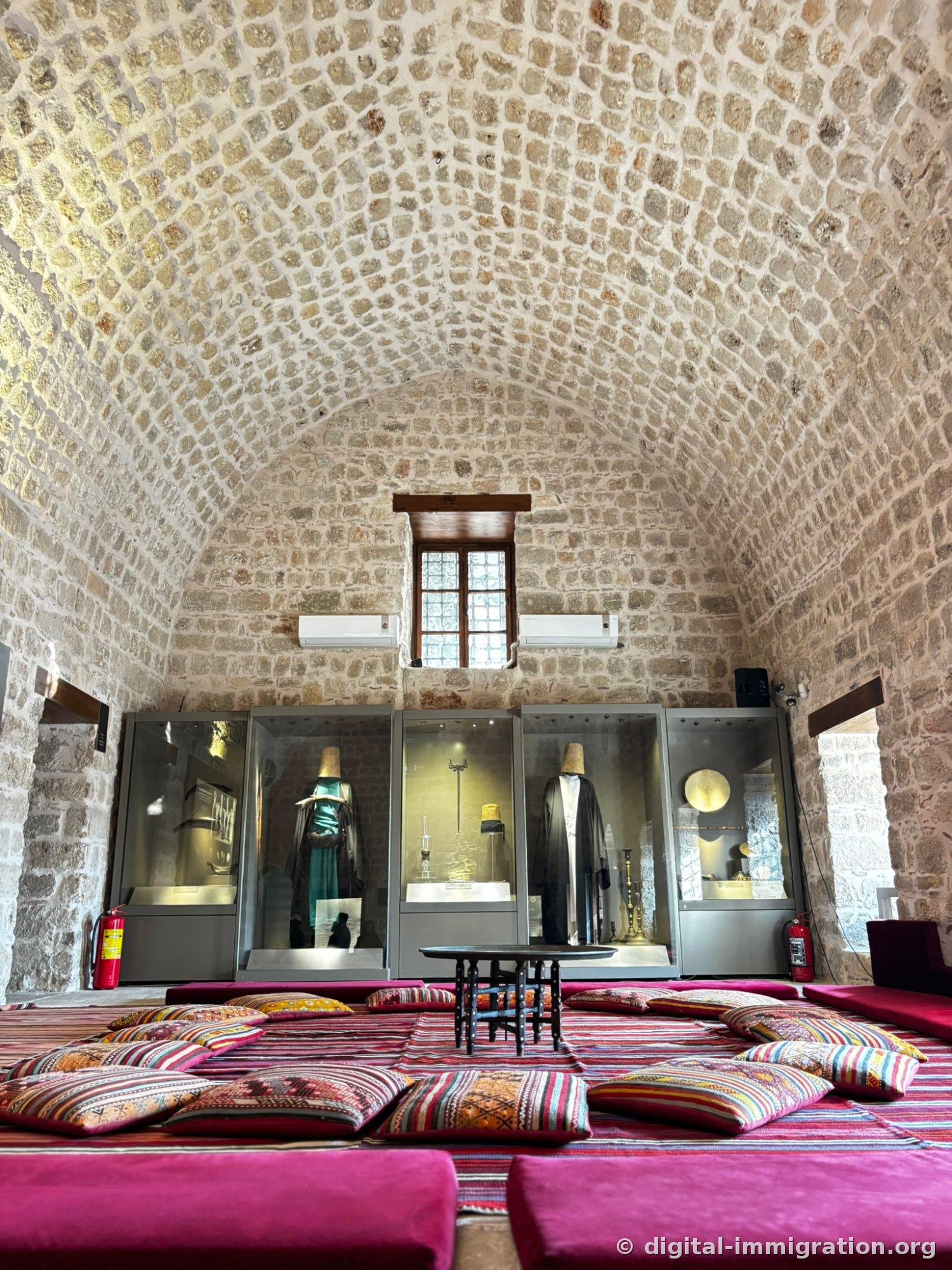 Haut plafond voûté en pierre sous lequel se trouvent cinq vitrines en verre avec chapeaux sikke et manteau noir, le sol recouvert de tapis mauve et coussins kilim