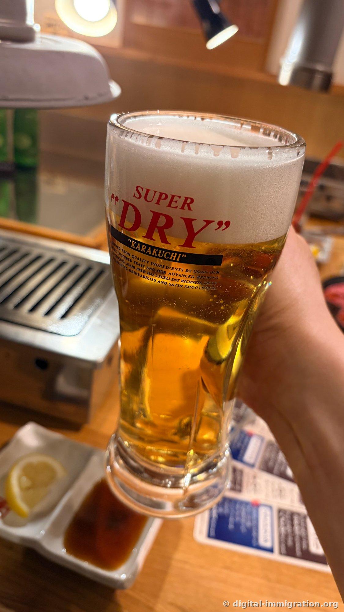 A hand holding an Asahi Super Dry draft beer glass. Behind it, a smokeless roaster and a white plate with a lemon wedge are visible