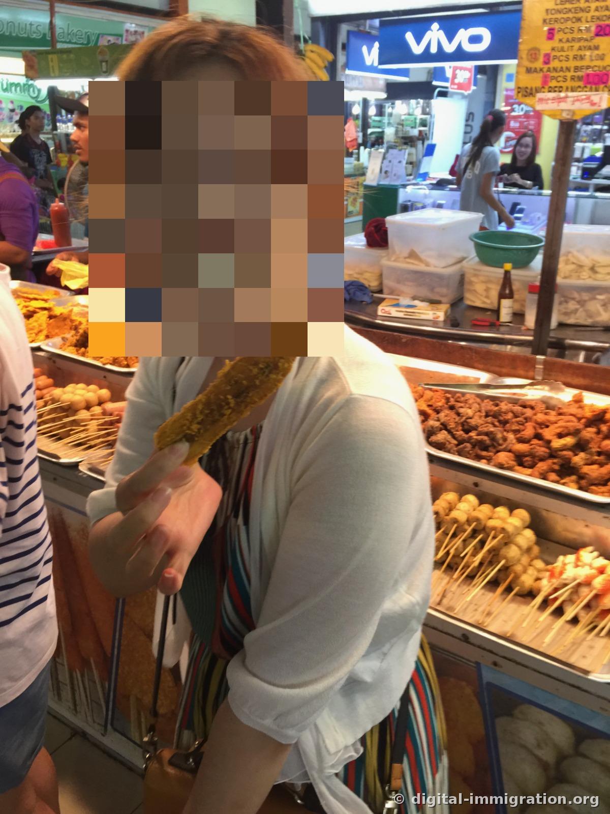 A person holding yellow fried food in front of a stall full of fried fish cake skewers and fried chicken skin, with a VIVO phone store sign visible behind