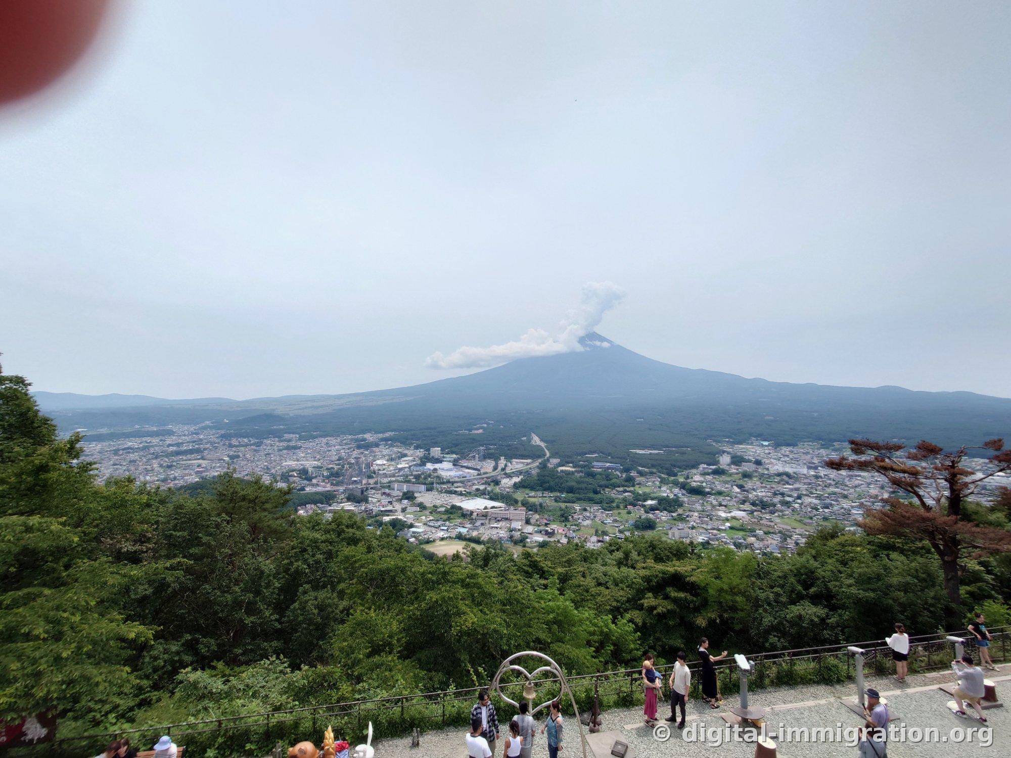 People gathered around the gravel ground and heart-shaped bell at the observation deck, with Mount Fuji billowing clouds and the Fujiyoshida town vista beyond