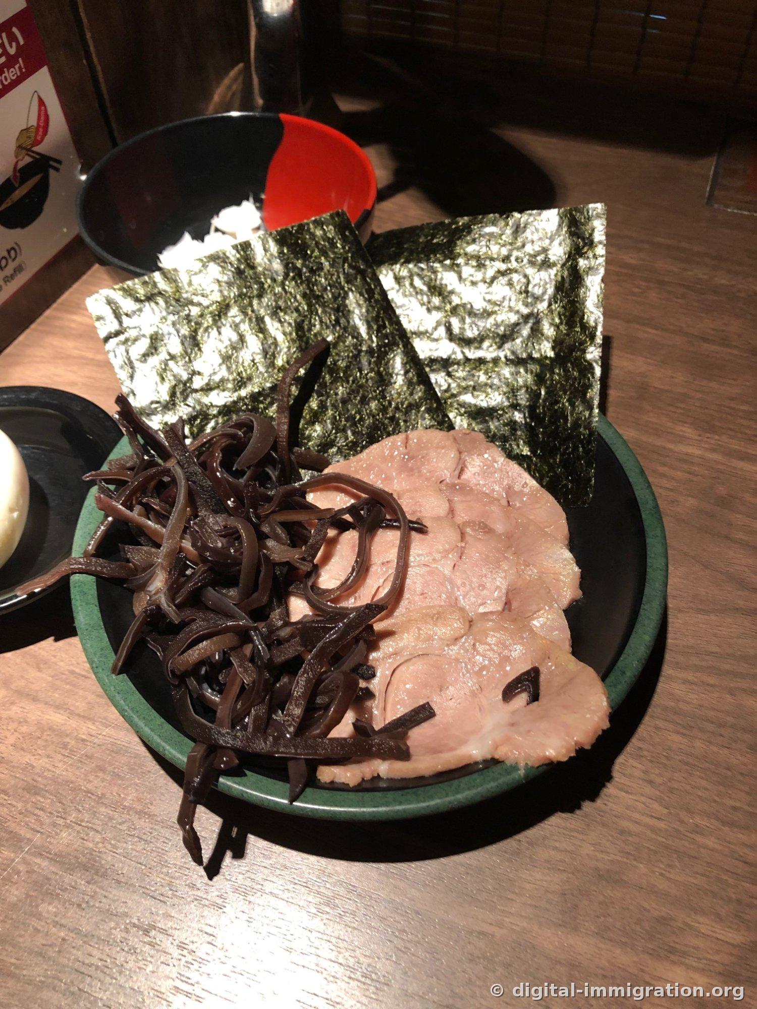 Sur une assiette à bord vert : plusieurs tranches de char siu et du kikurage, avec 2 feuilles de nori derrière, et un petit bol rougeâtre contenant l'œuf