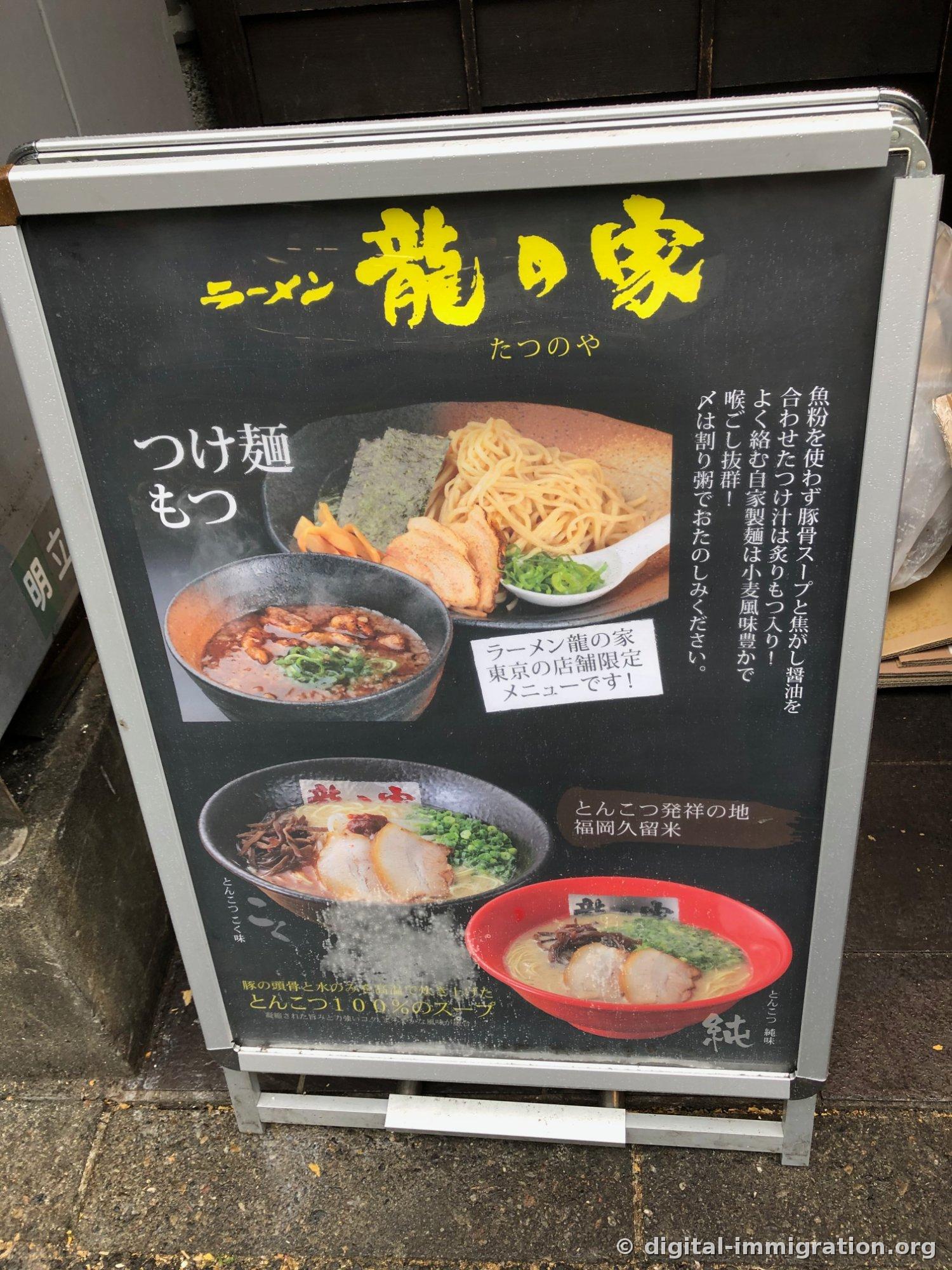 Un panneau en A noir devant le restaurant, montrant la photo de つけ麺もつ avec l'indication de menu exclusif à Tokyo en haut, et les photos du tonkotsu kokumi et junmai en bas