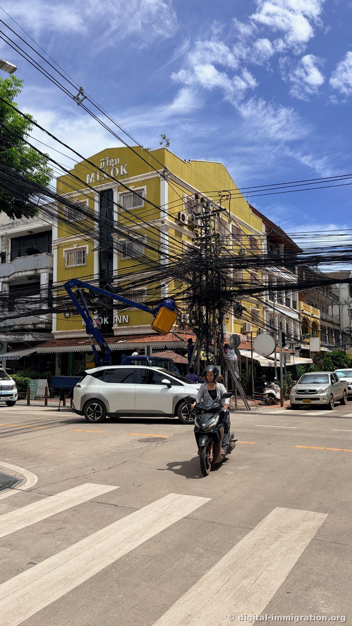 노란색 MIXOK INN 건물 앞 사거리, 거대한 전선 뭉치 전봇대에 파란 고소작업차 바스켓과 사다리를 타고 올라간 작업자들, 앞으로 지나가는 스쿠터