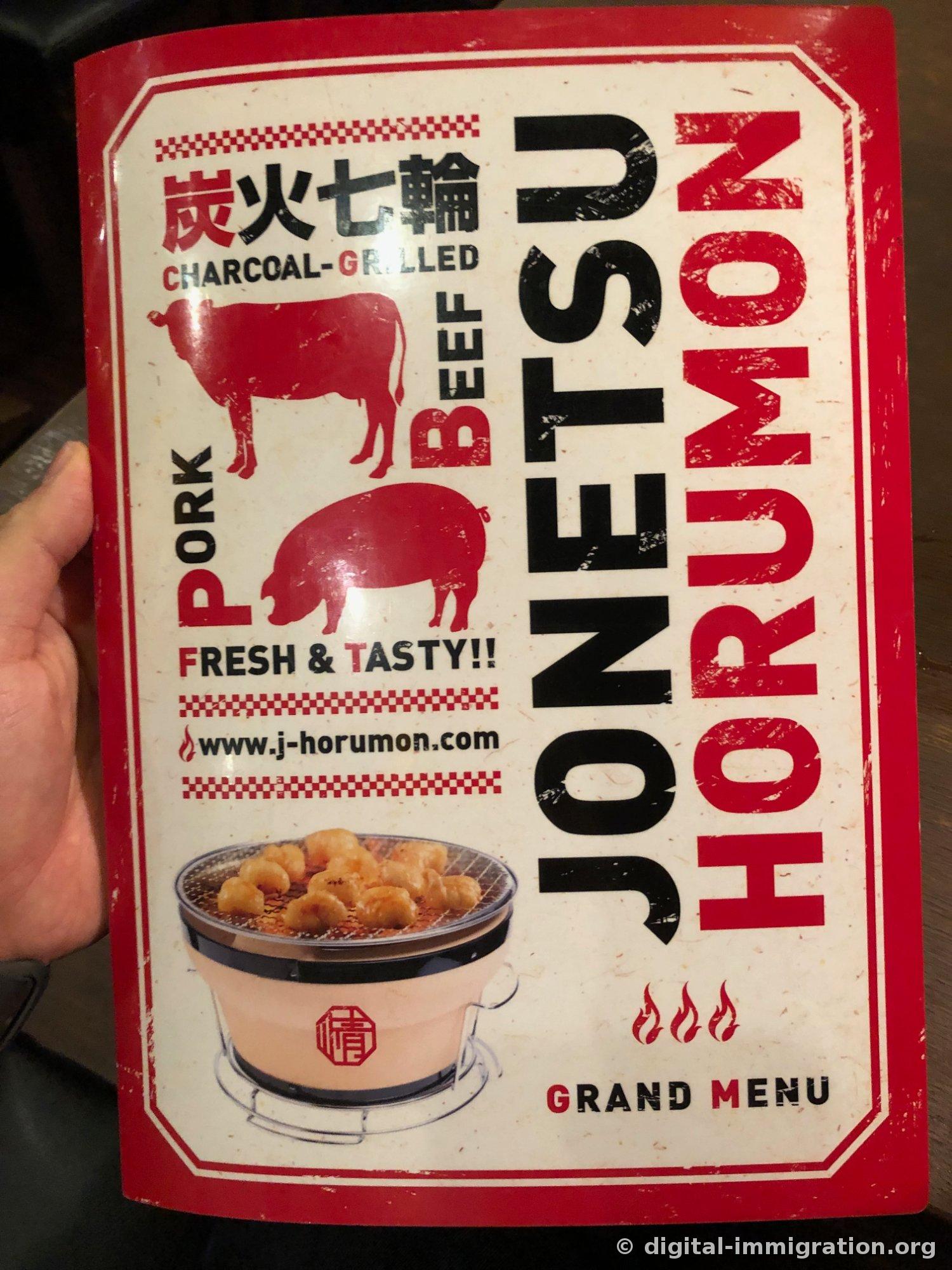 A hand holding a grand menu with a red cover featuring drawings of a cow and pig, the Jonetsu Horumon logo, and a shichirin grill photograph